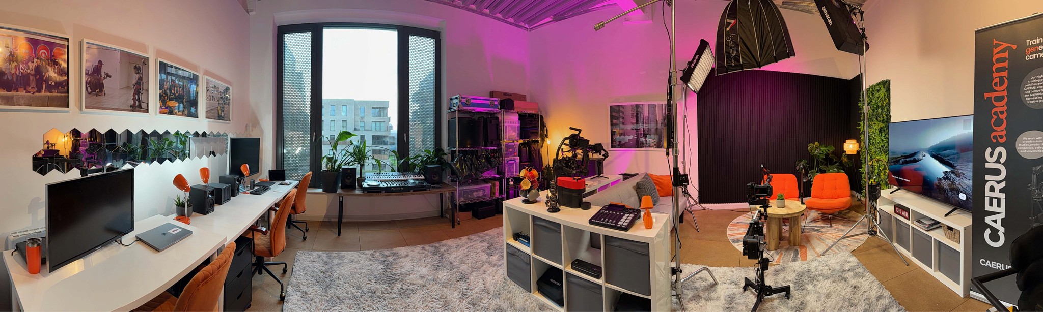 Image of our studio in Hackney Wick showing our editing suite, production kit and podcast studio. The studio has orange elements including seating and lamps, white tables, a screening area, podcast studio, lots of plants and a large window that lights it along with some warm lighting from pink RGB lights.