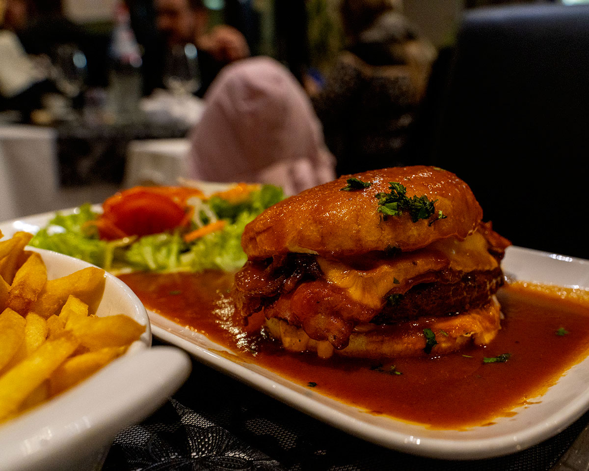 Hamburger maison nappé de sauce Francesinha, servi au Restaurant Miragem à Pétange, Luxembourg, accompagné de frites croustillantes et d'une salade fraîche. Une fusion gourmande entre le burger classique et la cuisine portugaise.