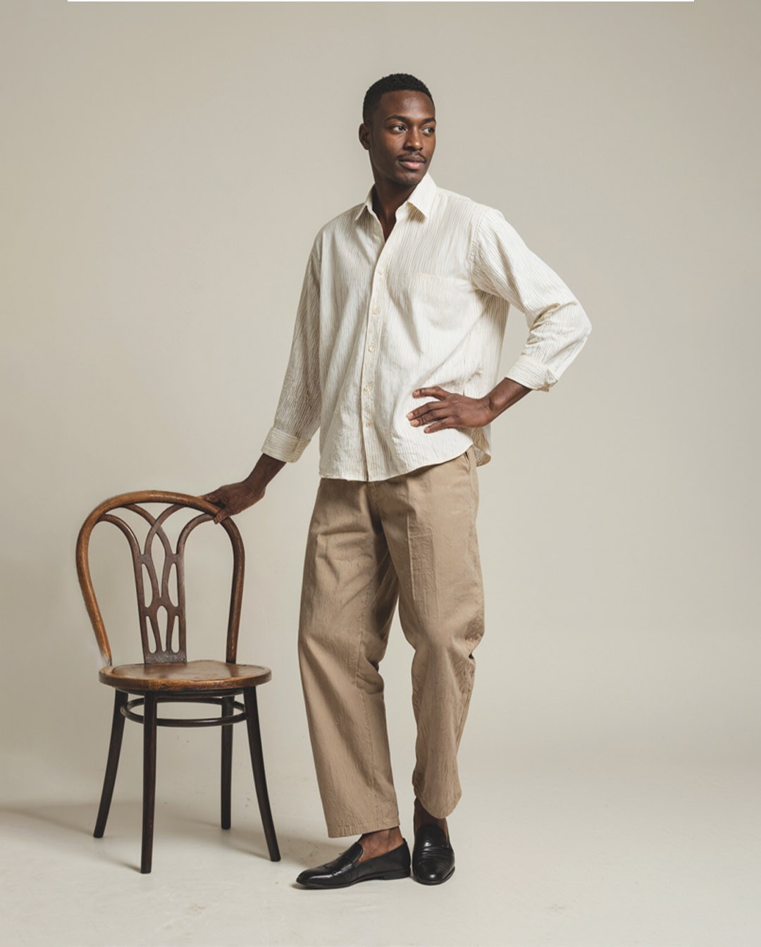 Stylish man standing next to a wooden chair wearing a cream button-up shirt, beige pants, and black loafers, minimalist studio fashion look
