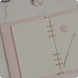 Open blush pink planner with a gold ruler on a light surface.