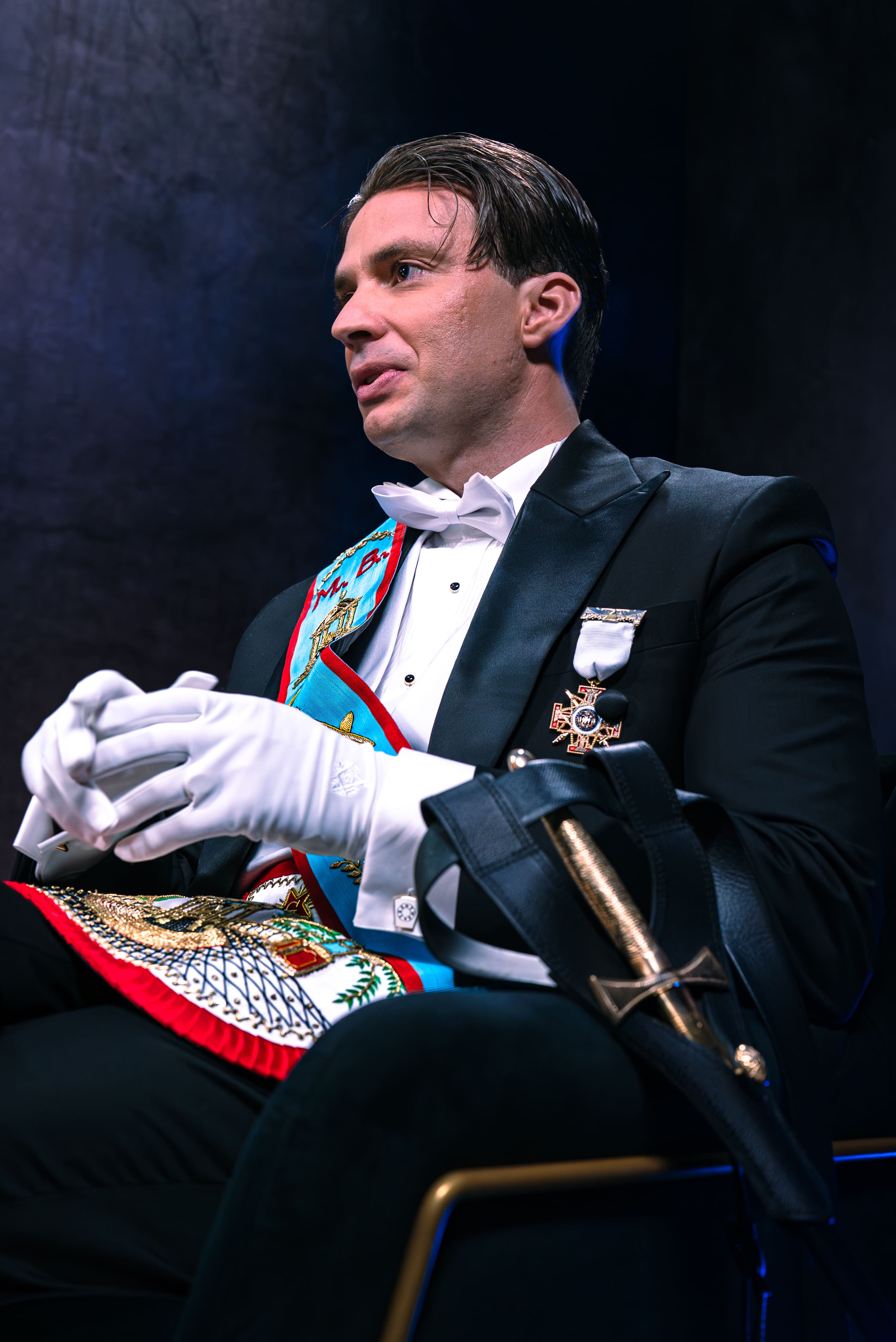 Photographie de portrait professionnelle d'un homme en tenue de Franc-Maçonnerie (smoking, gants blancs, écharpe d'officier colorée) interviewé dans le studio Legend. L'homme est assis, le regard dirigé hors champ, dans une posture d'écoute attentive. Une épée cérémonielle dans son fourreau est posée sur ses genoux. Le costume et les insignes sont très détaillés. L'arrière-plan est sombre, éclairé par des touches de lumière bleue. Ce portrait, optimisé par Nux-Studio, illustre la production photo pour des interviews formelles, des sujets d'enquête et de société, mettant en évidence l'éclairage de studio et la direction artistique.
