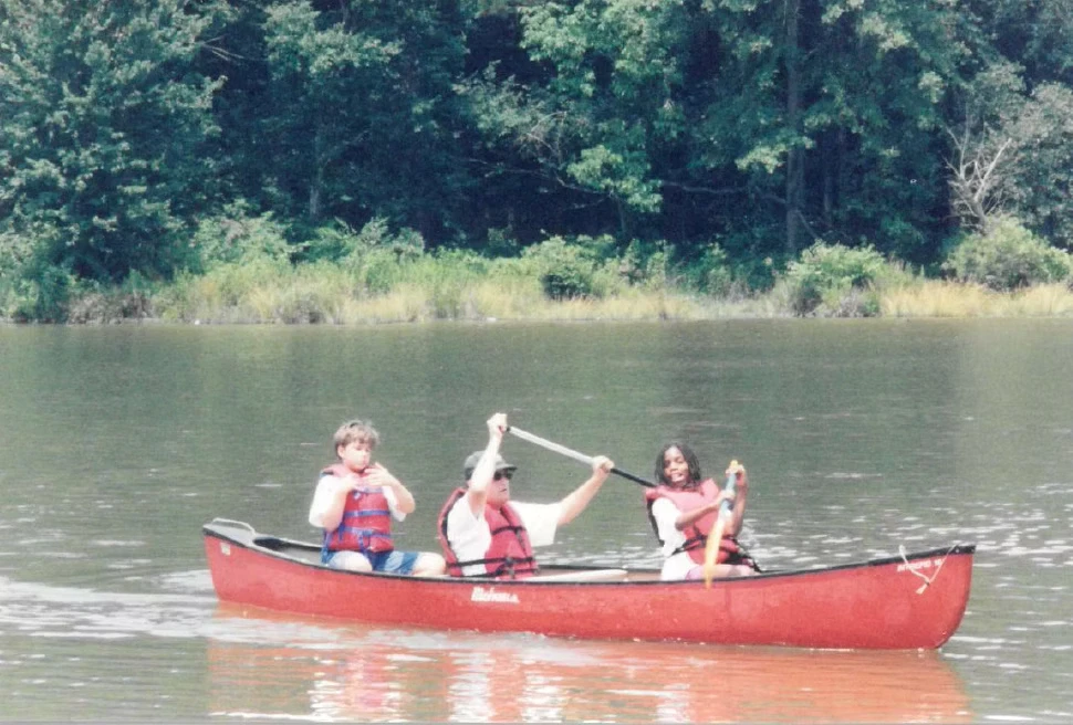 Three people in a red canoe on a calm lake, paddling and enjoying a sunny day. Lush green trees line the shore.