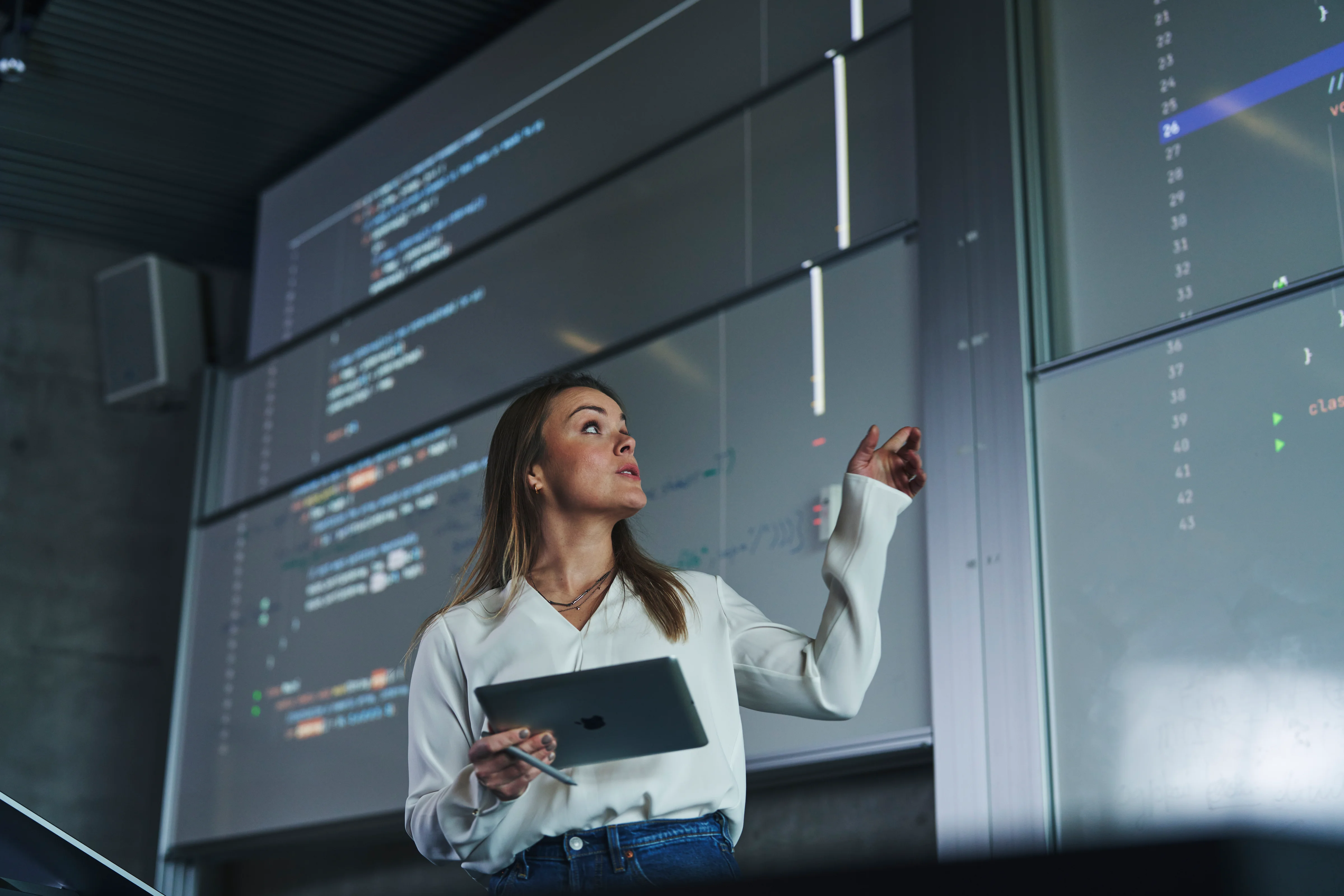 A woman holding a tablet gestures toward large digital displays during a presentation, illustrating Iceland’s modern, tech-driven work environment.