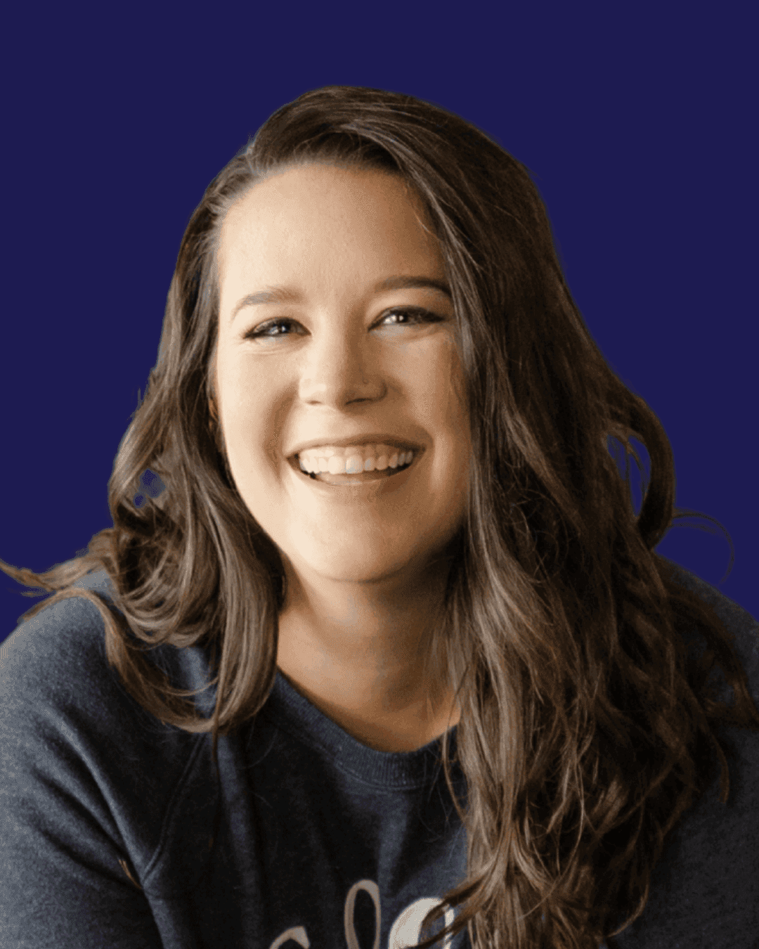 A person with long, wavy brown hair and a joyful expression is smiling against a solid deep blue background, wearing a dark sweater.