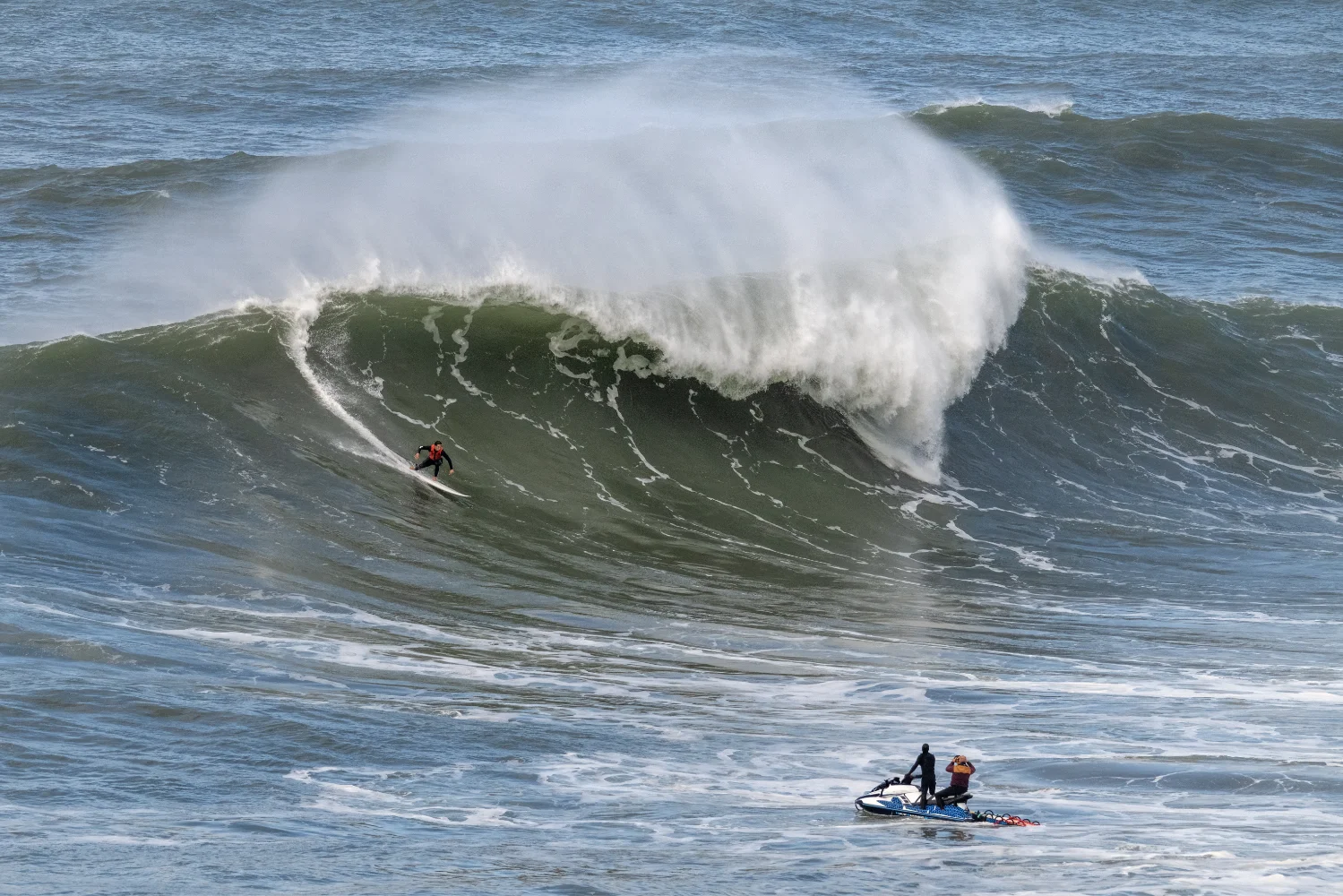 Surfer riding a large wave with Jet Ski support nearby.