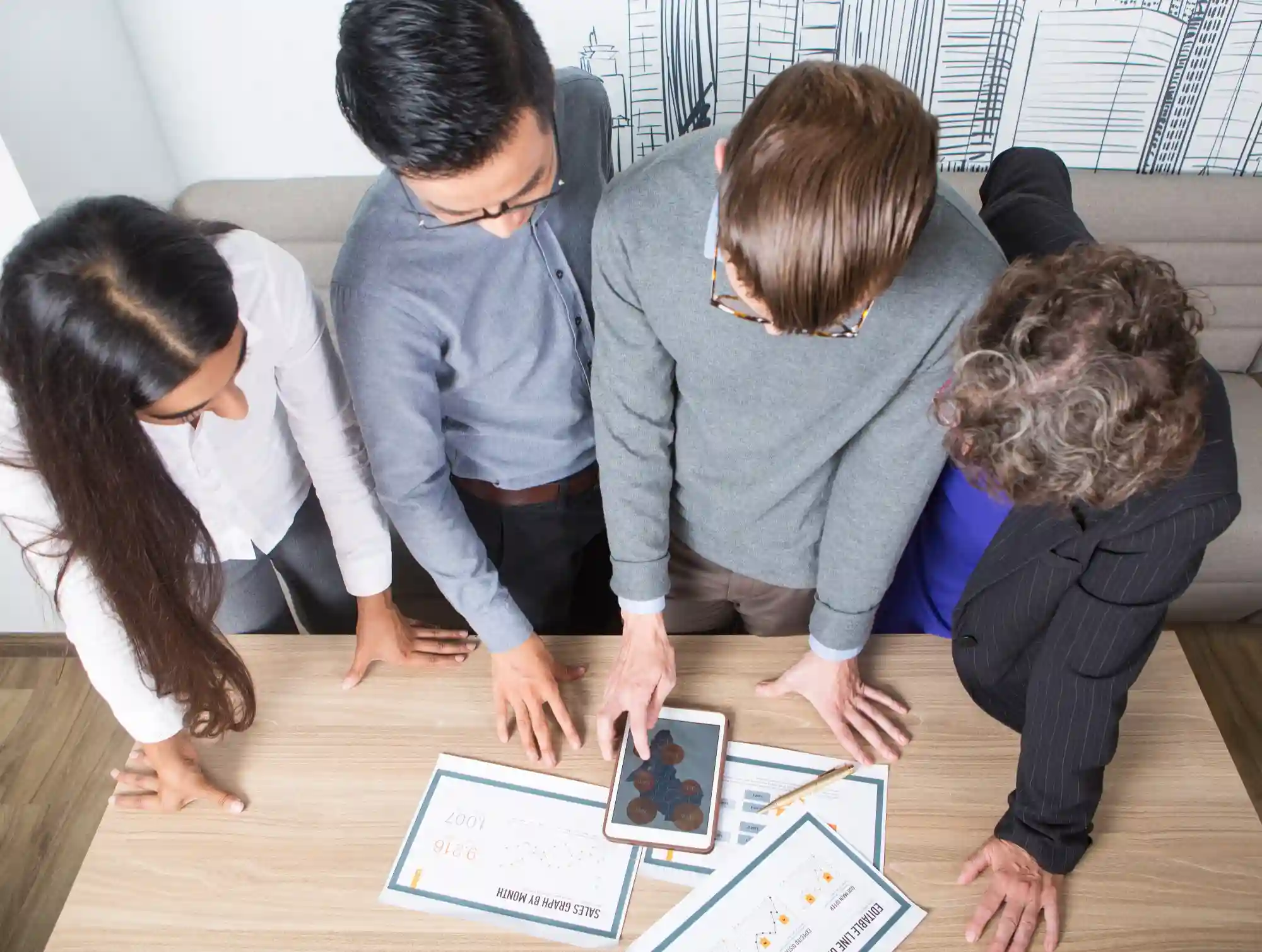 A diverse team of professionals gathers around a desk to review data charts and analytics on a digital tablet.