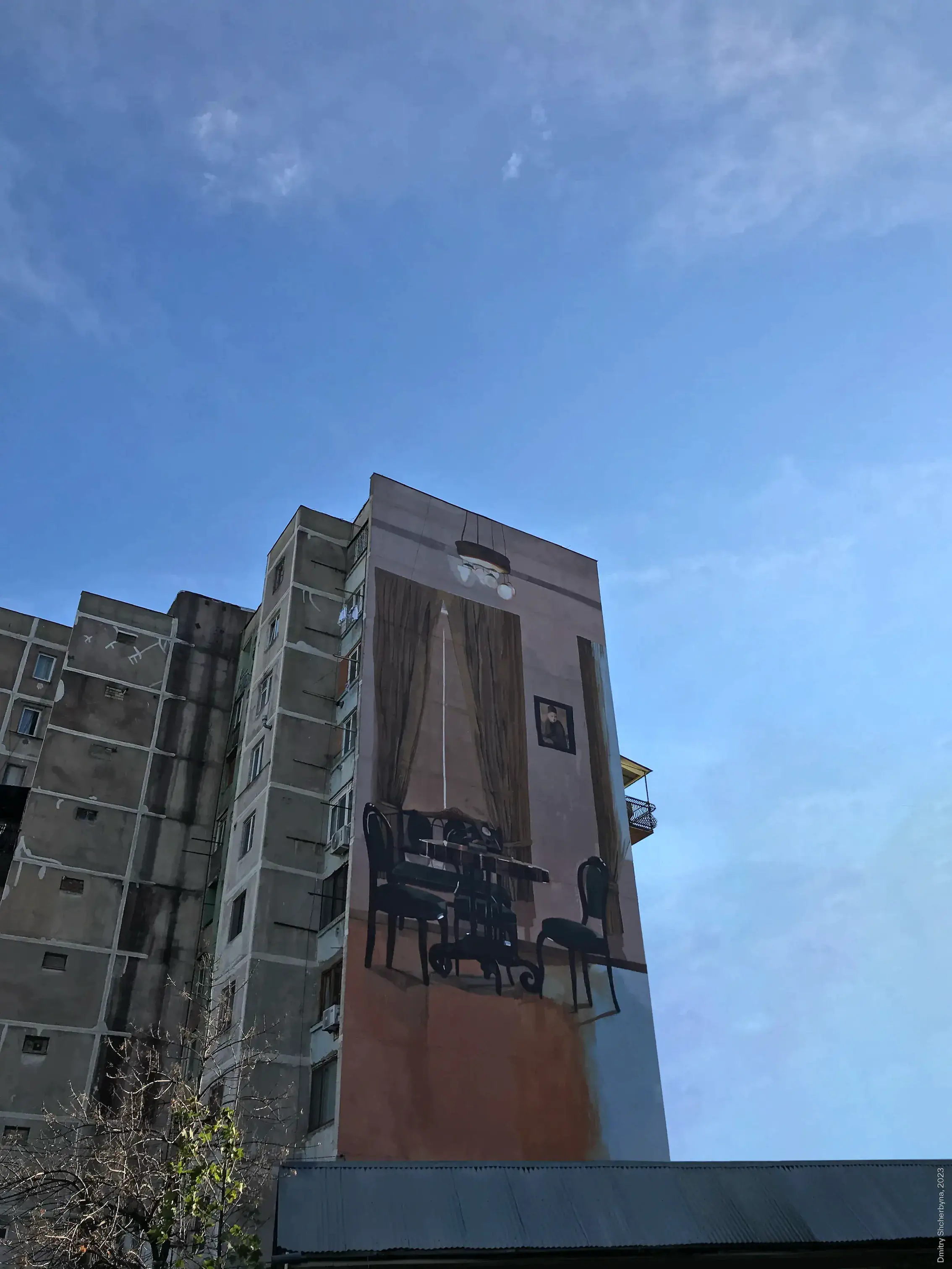 A mural on the side of a high-rise apartment building depicts a traditional interior room with dark wooden furniture, a table with chairs, long curtains flanking a bright window, a framed portrait on the wall, and a hanging light fixture. The painting creates the illusion of a room on the building's exterior.