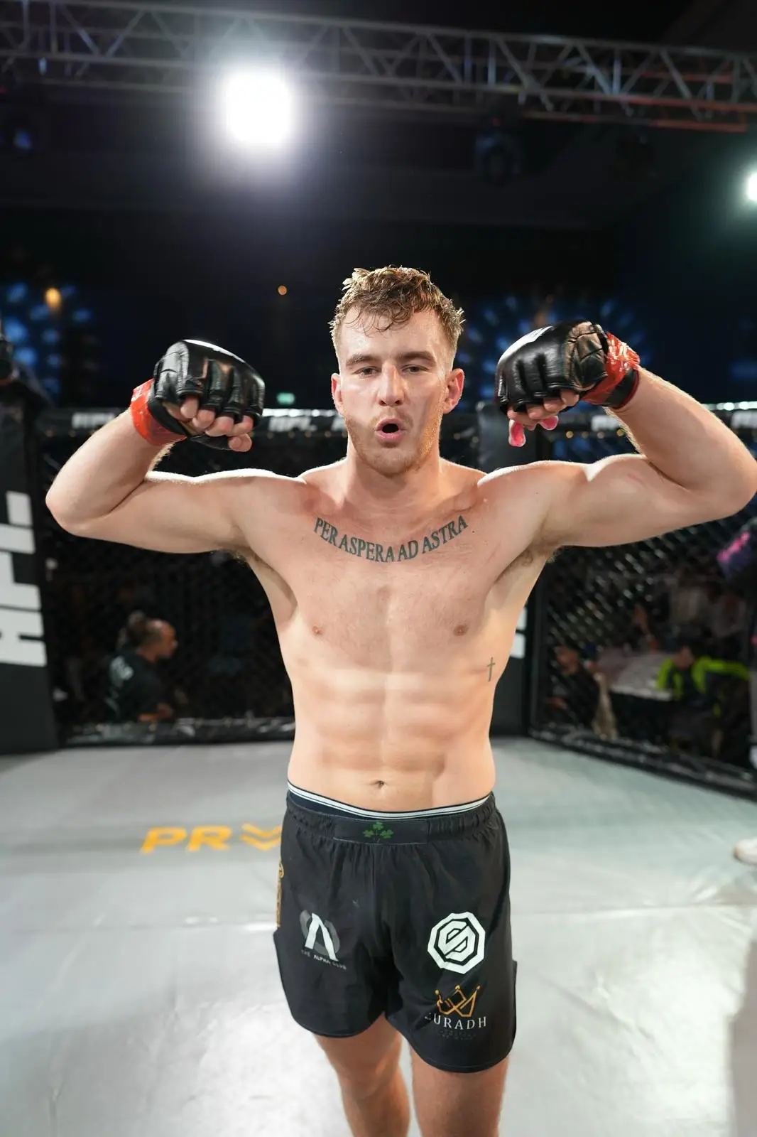 Paul Buckley flexing inside an MMA cage, wearing gloves — MMA and strength specialist at The Alpha Club.