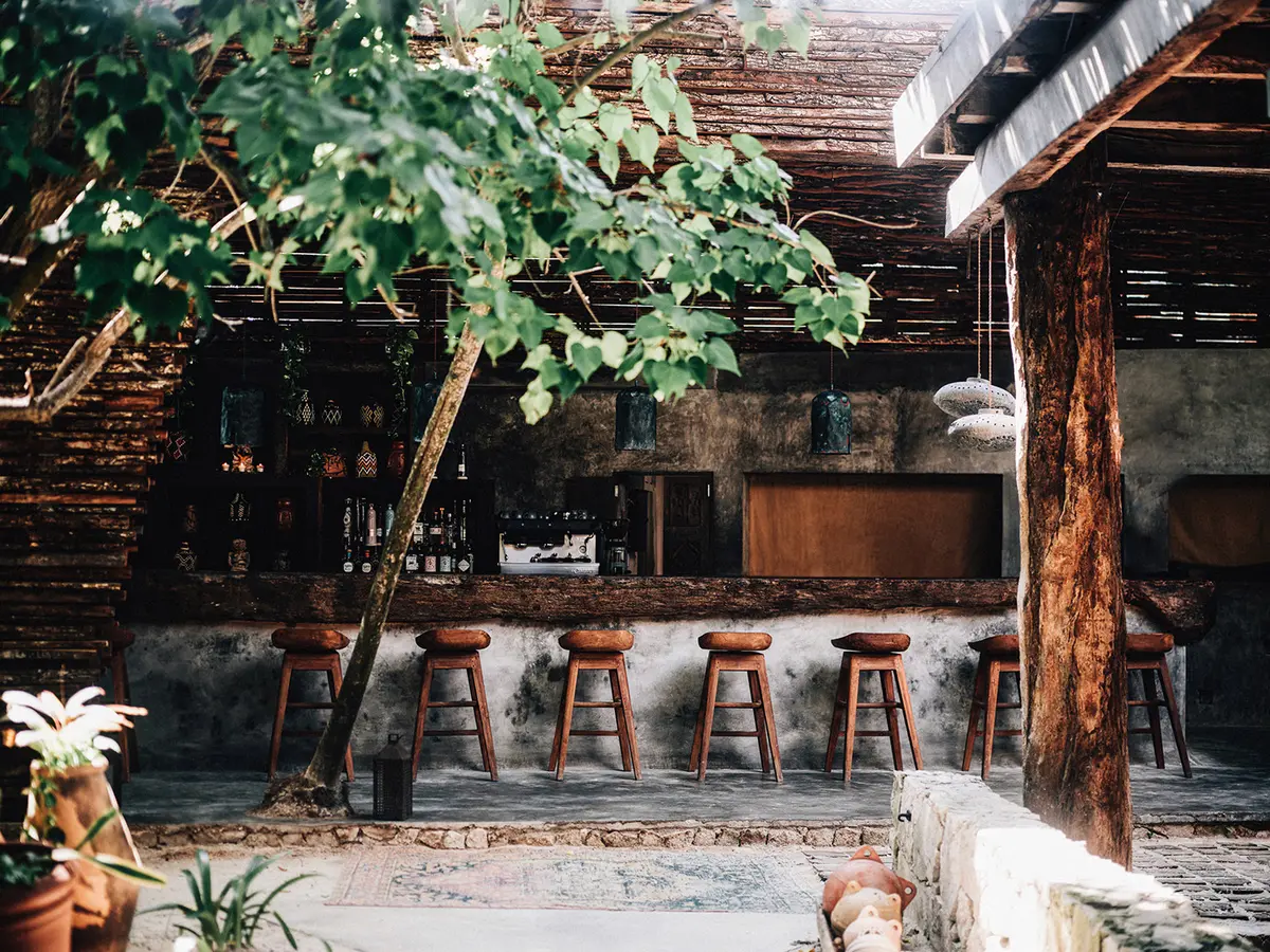 View of the bar at Macondo restaurant in Nômade Tulum, Mexico, featuring counter and seating for guests, small trees, and a Mediterranean-style ceiling made of tree trunks.