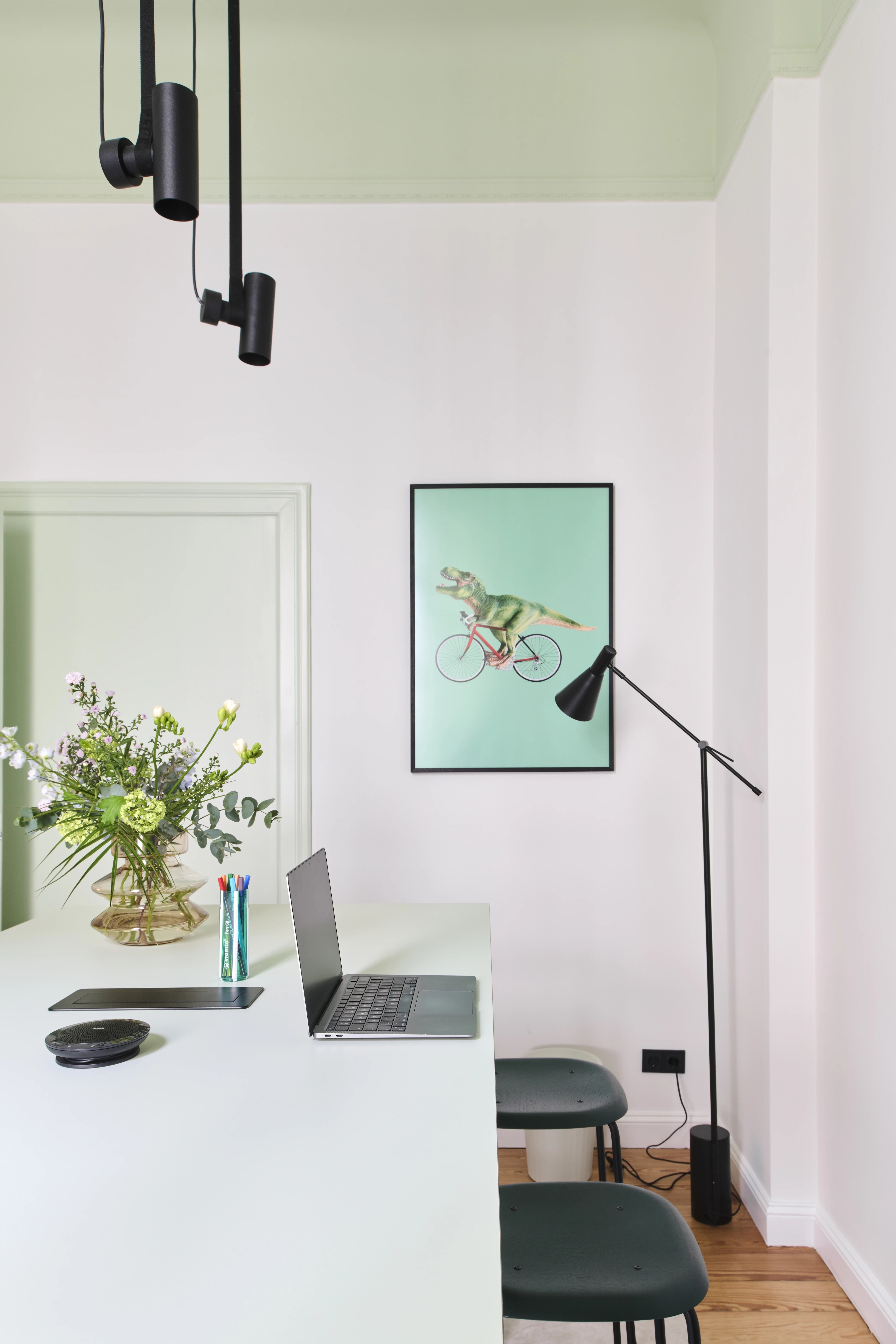 Workspace at INTERMATE BERLIN with a mint-green desk with laptop and flower bouquet, dark green stools, a black floor lamp, a dinosaur-on-bike poster and a pale green ceiling.