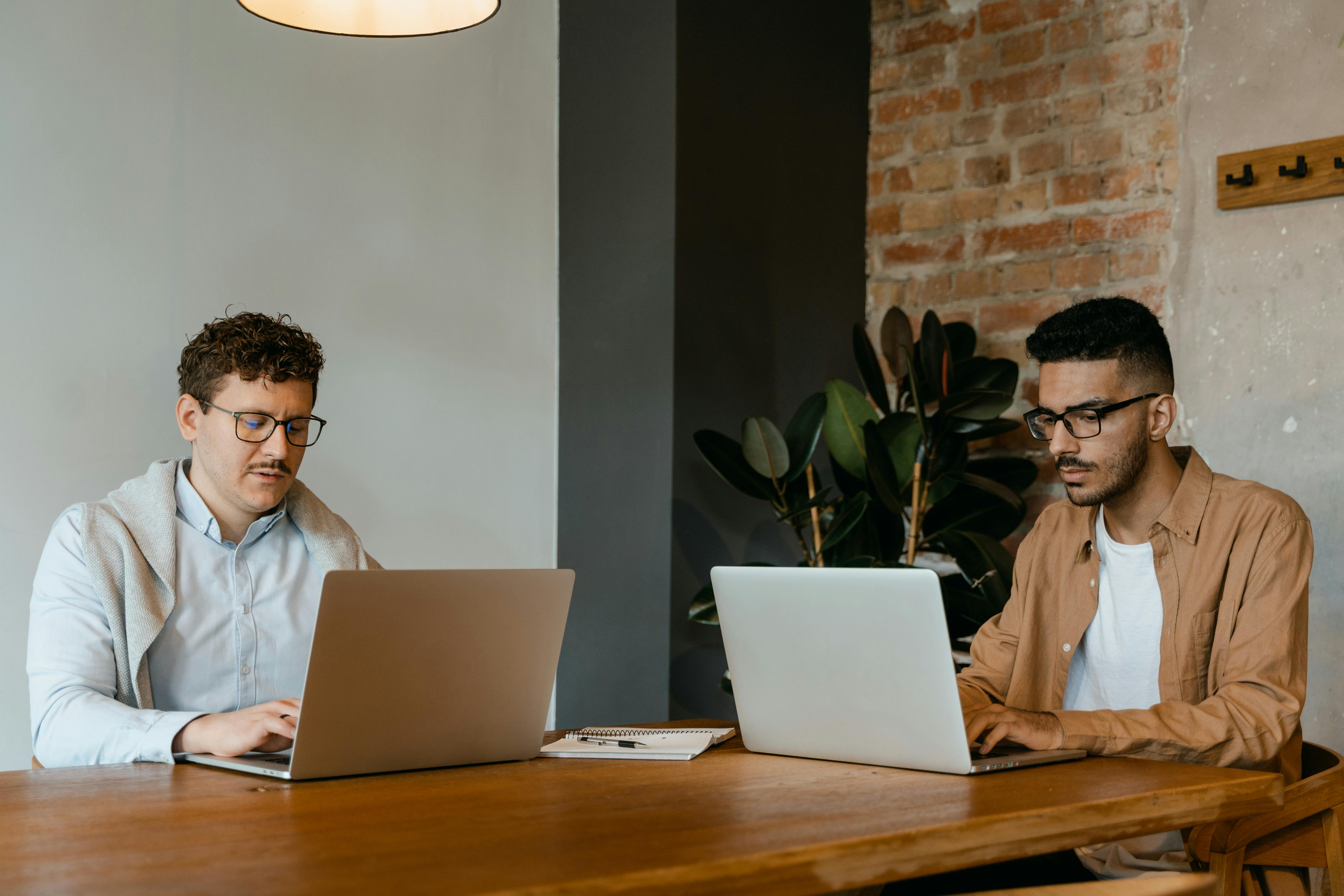 Two young men are sitting at a table, working on laptops in a modern, well-lit office space.