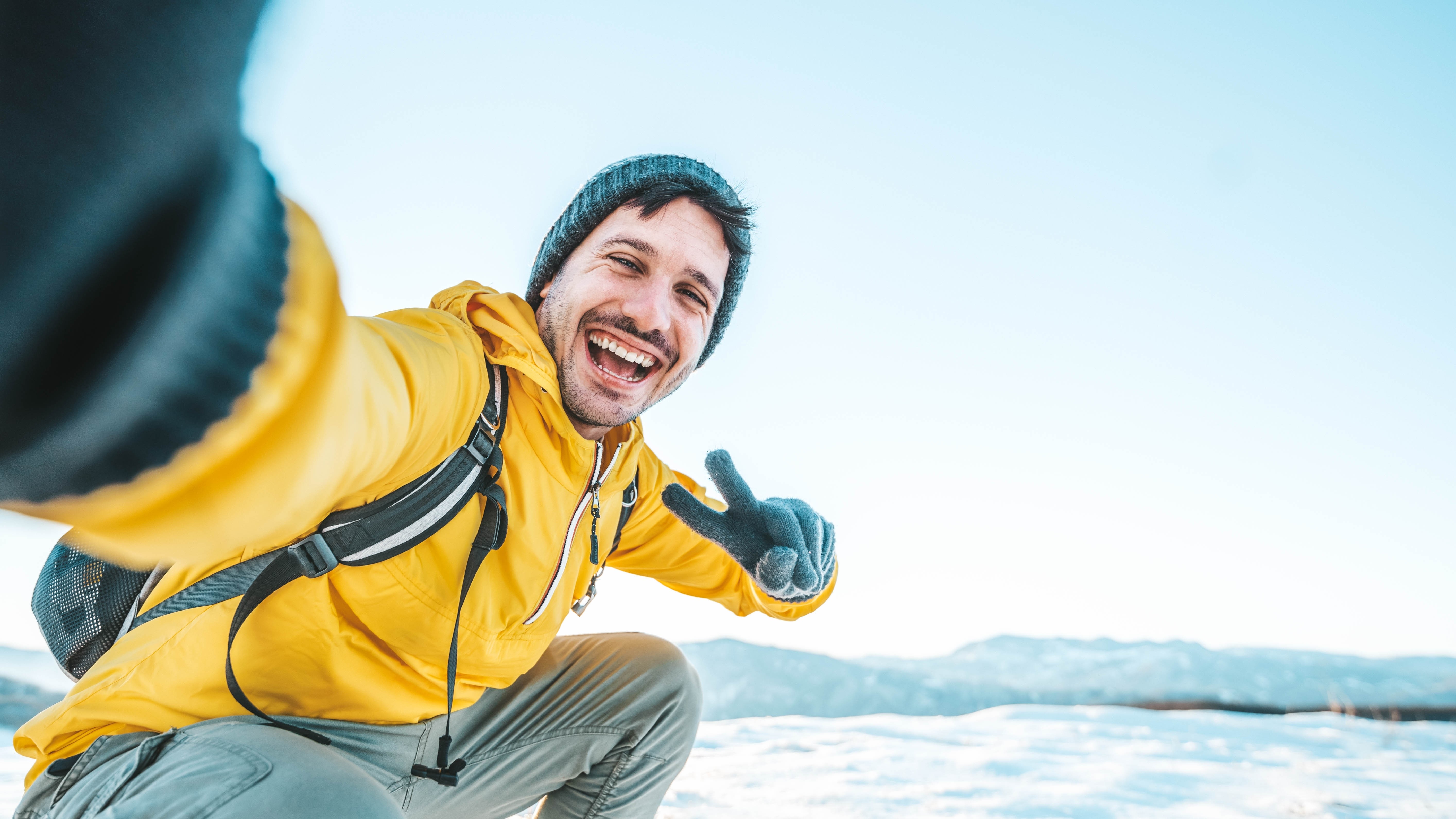 A person in a yellow jacket taking a selfie and smiling with peace fingers on a snowy mountain landscape.