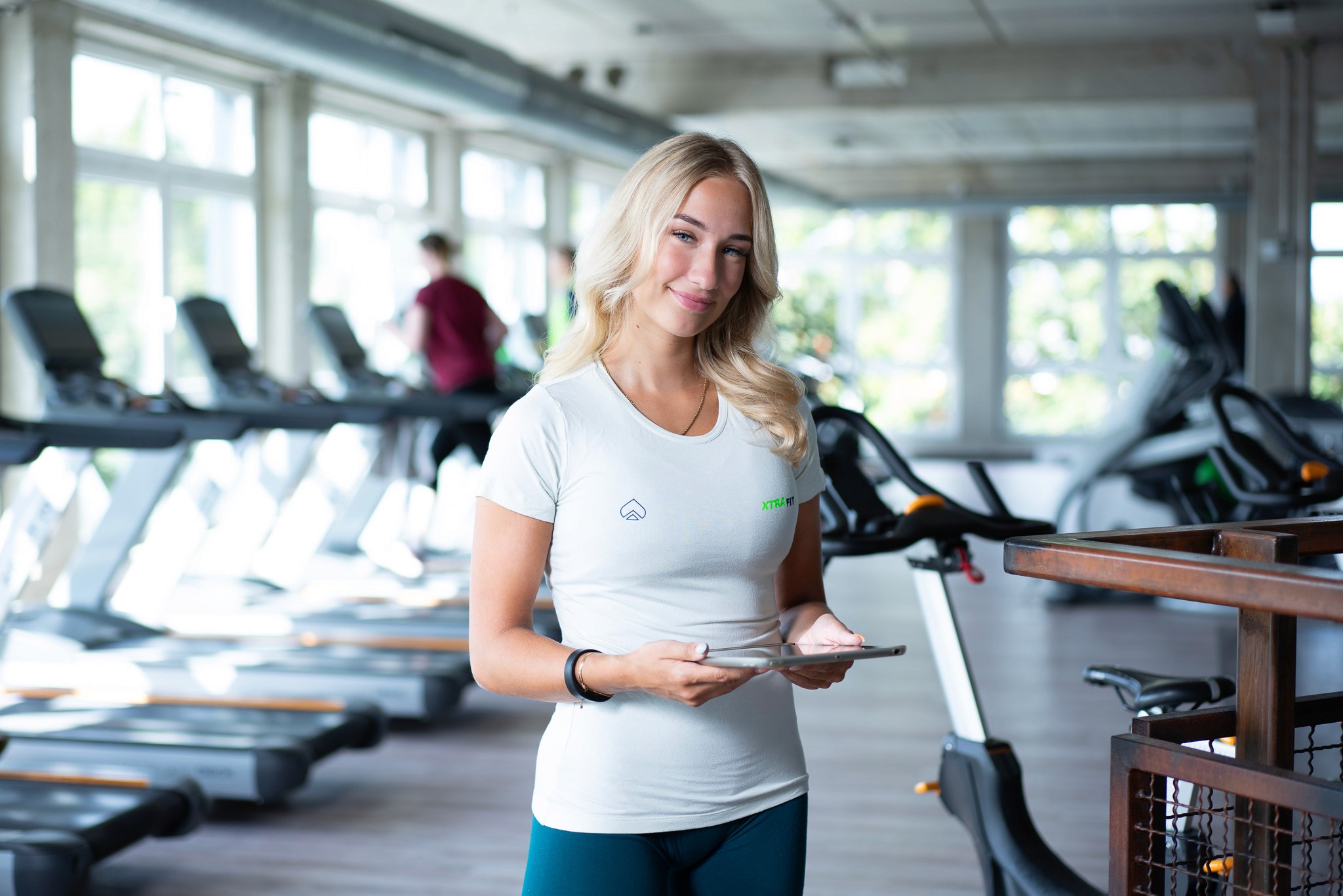 Eine XTRAFIT-Trainerin steht mit einem Tabeltcomputer in der Hand im Cardio-Bereich eines XTRAFIT-Studios.