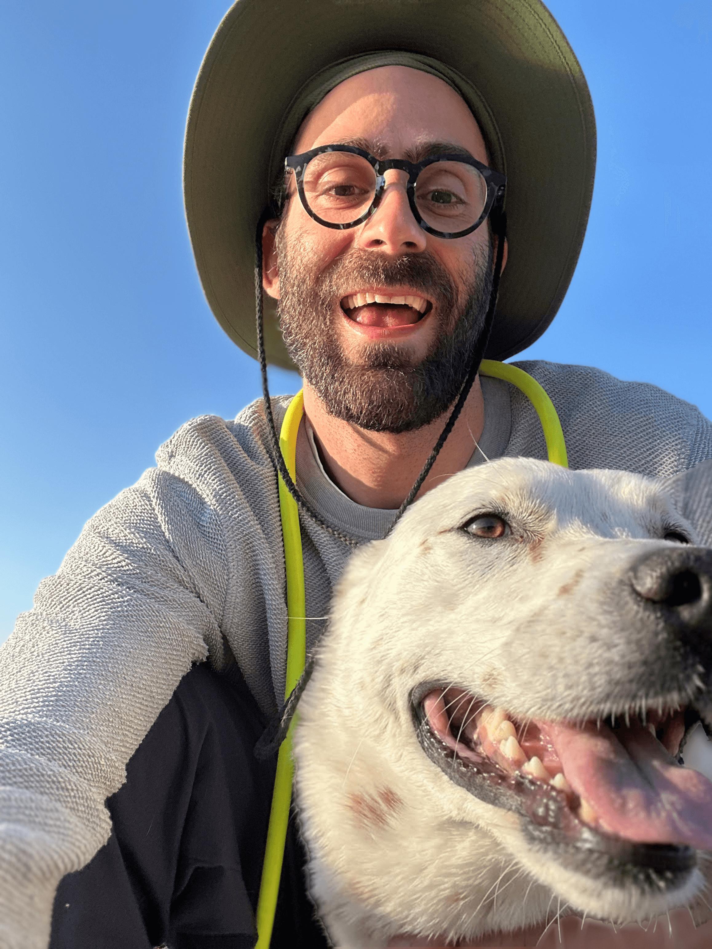 Evan in a sun hat and glasses, smiling, with his white dog Willow beside him under a blue sky.