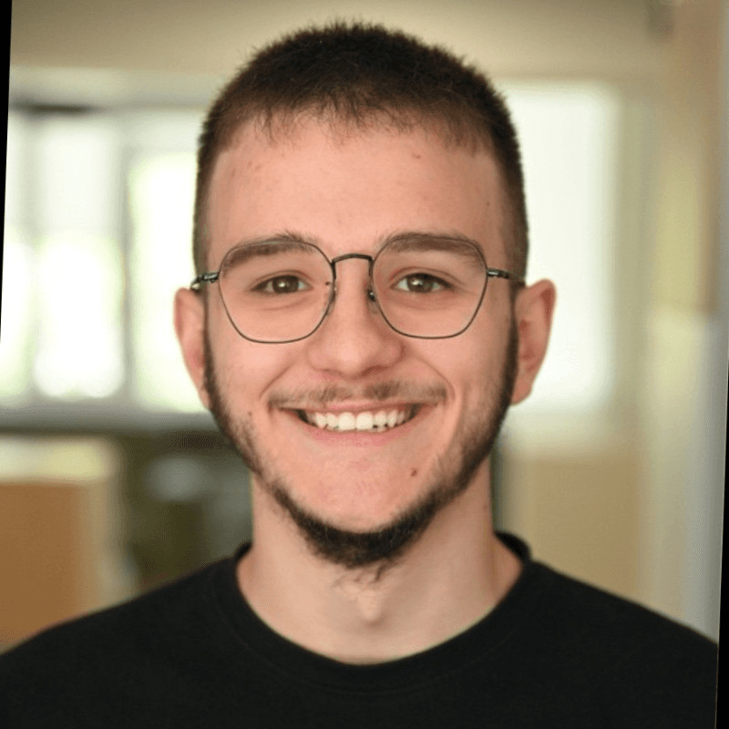 A smiling person with short hair and glasses, wearing a black shirt, stands in a bright indoor setting.