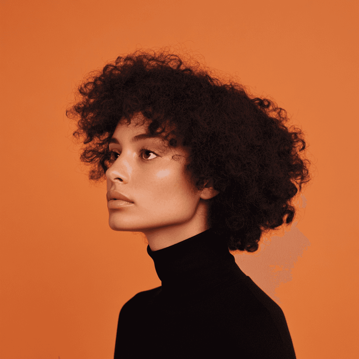 Jeune femme à la peau mate et aux cheveux bouclés, vêtue d'un col roulé noir, regardant pensivement vers la droite sur un fond orange uni.