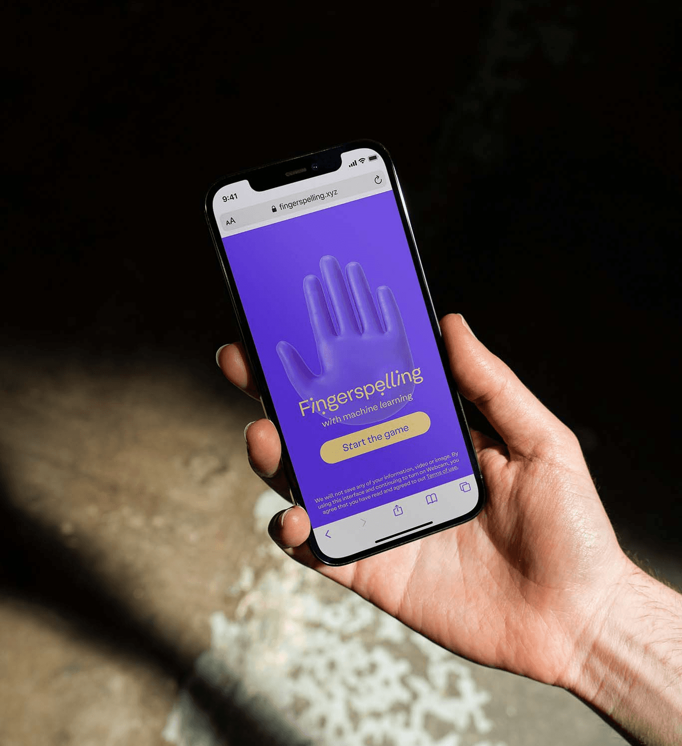 A person holds a smartphone in a sunlight-dappled setting, displaying the "Fingerspelling with machine learning" home screen. This lifestyle mockup emphasizes the global accessibility and portability of the software designed by the branding studio.