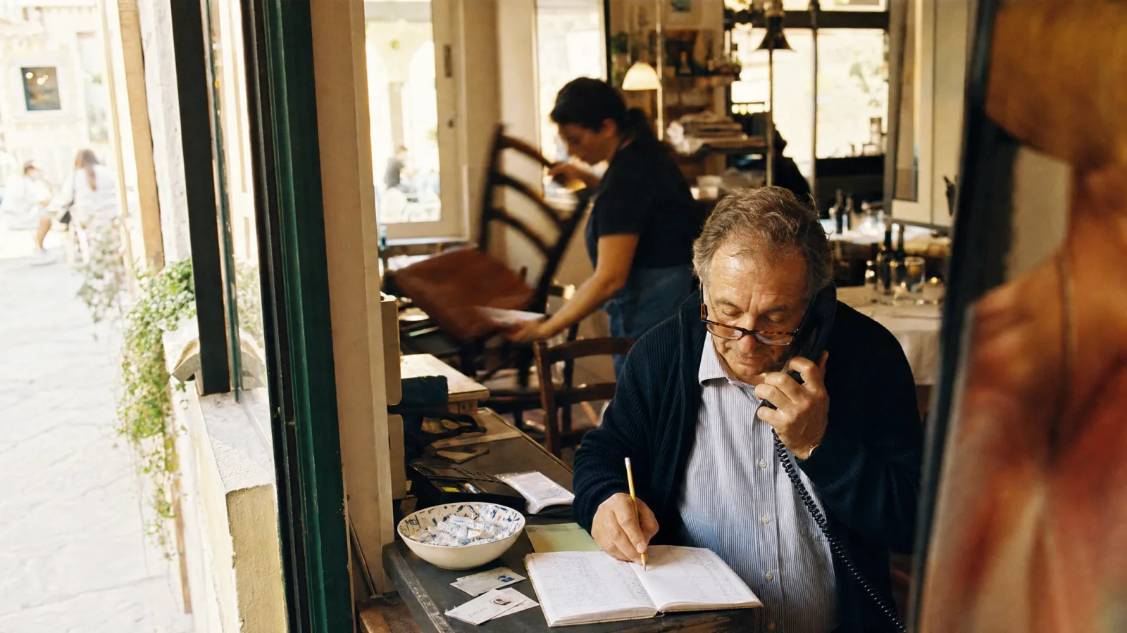 Proprietario prende una prenotazione al telefono scrivendola a matita sull'agenda della trattoria