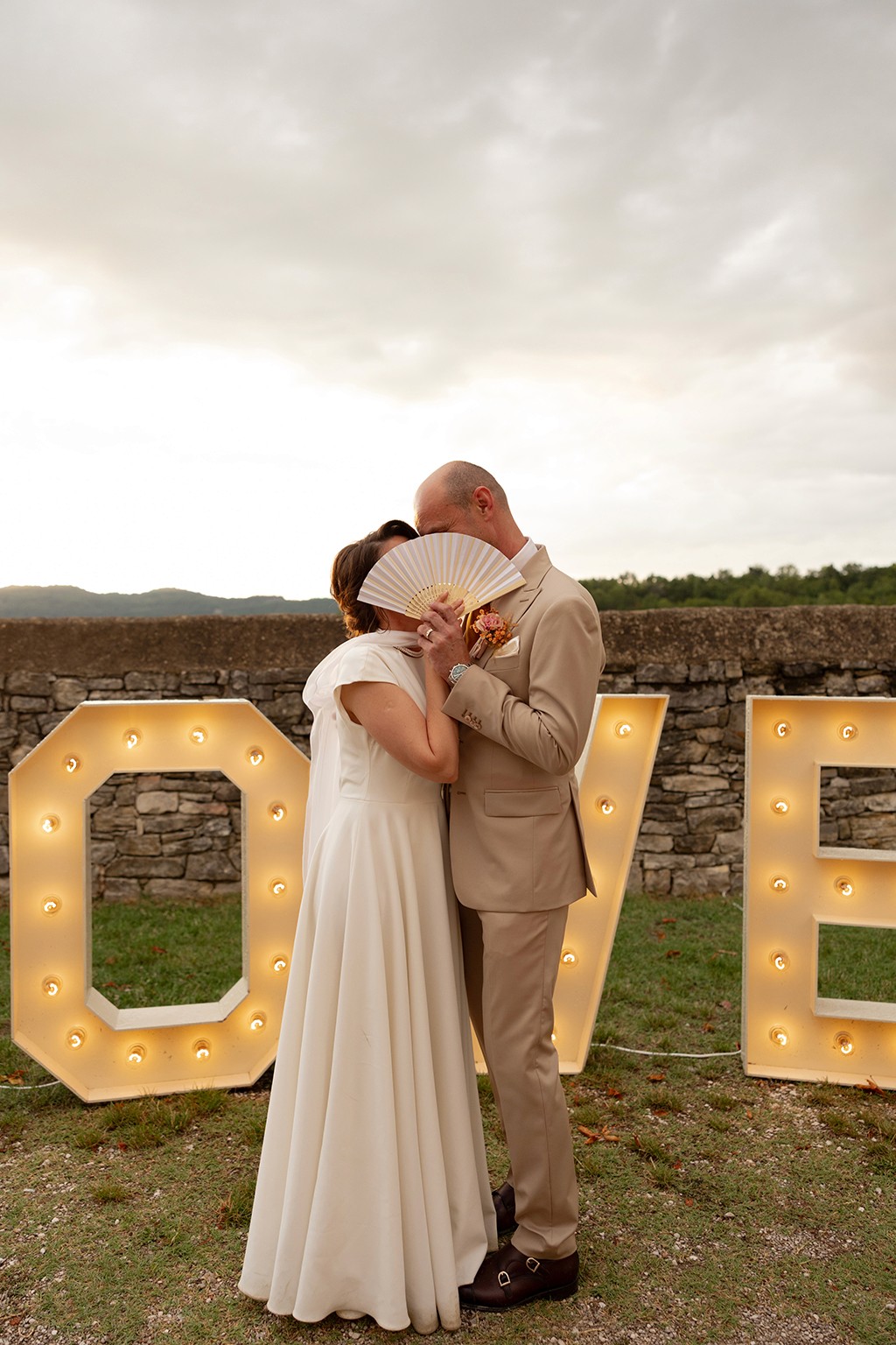 Married couple posing for a photo in front of an old wall, holding a fan over their faces, in front of a LOVE sign