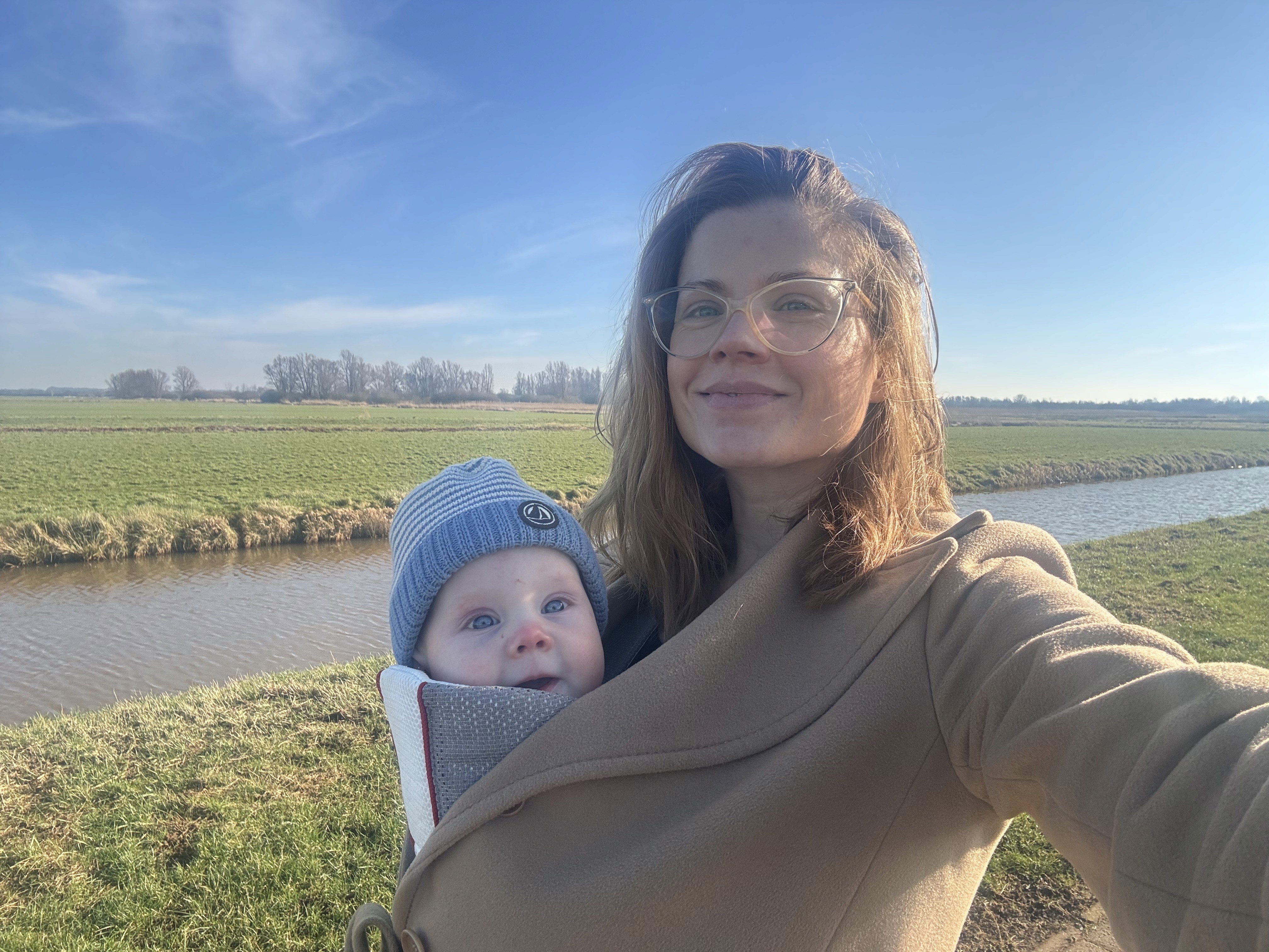 Tosca de Jong on a walk through the Dutch polder landscape with her baby in a beige coat and carrier, standing by a canal under a bright blue sky.