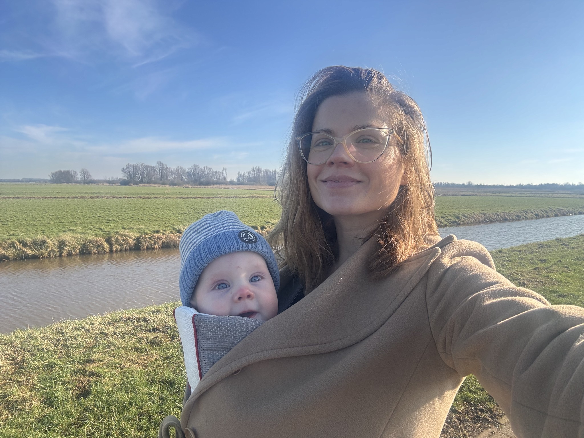 Tosca de Jong on a walk through the Dutch polder landscape with her baby in a beige coat and carrier, standing by a canal under a bright blue sky.