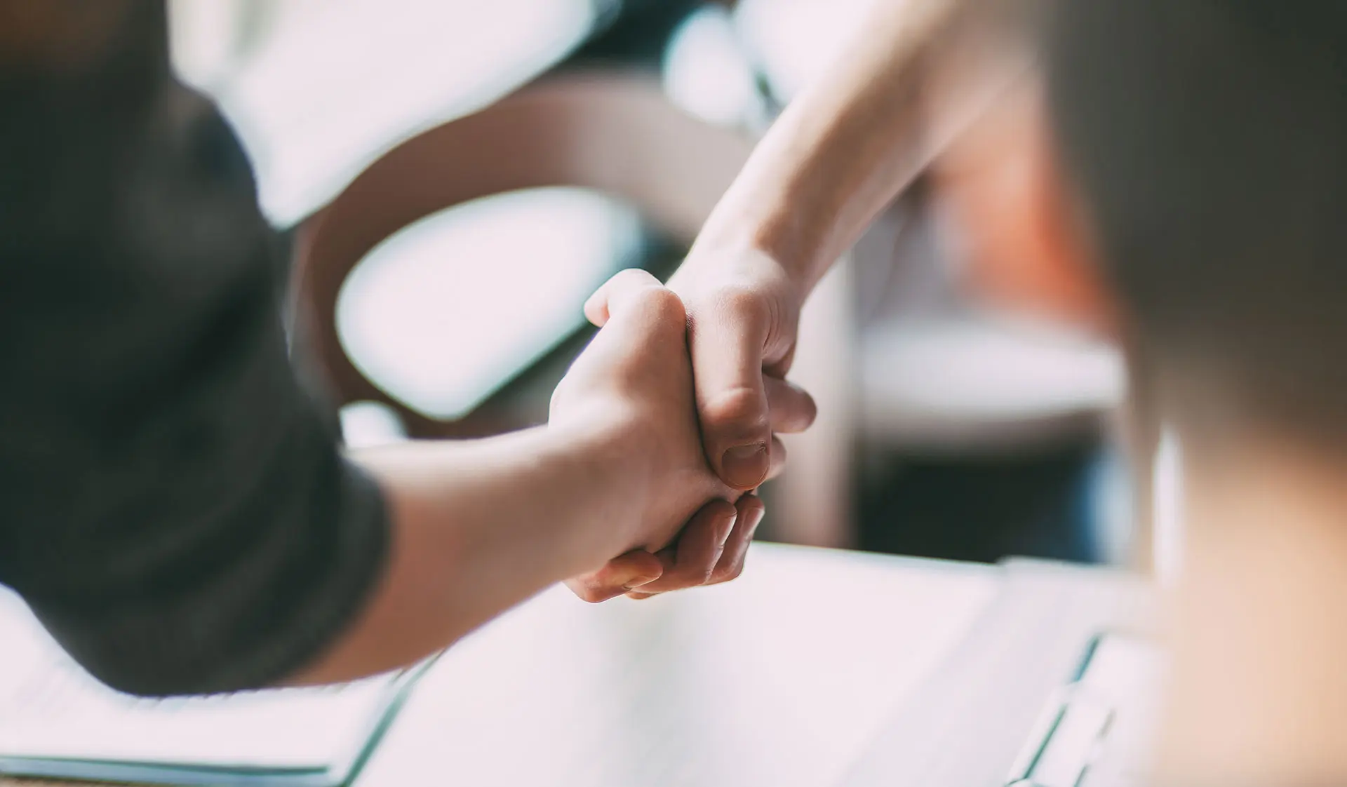 Close-up of two professionals shaking hands across a desk with an open laptop, signalling a business agreement