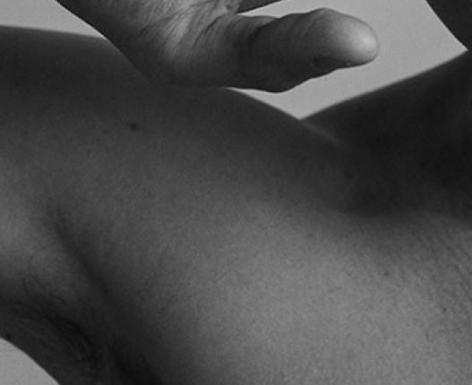 Black and white close-up of a person’s bare shoulder and raised arm in soft studio lighting.