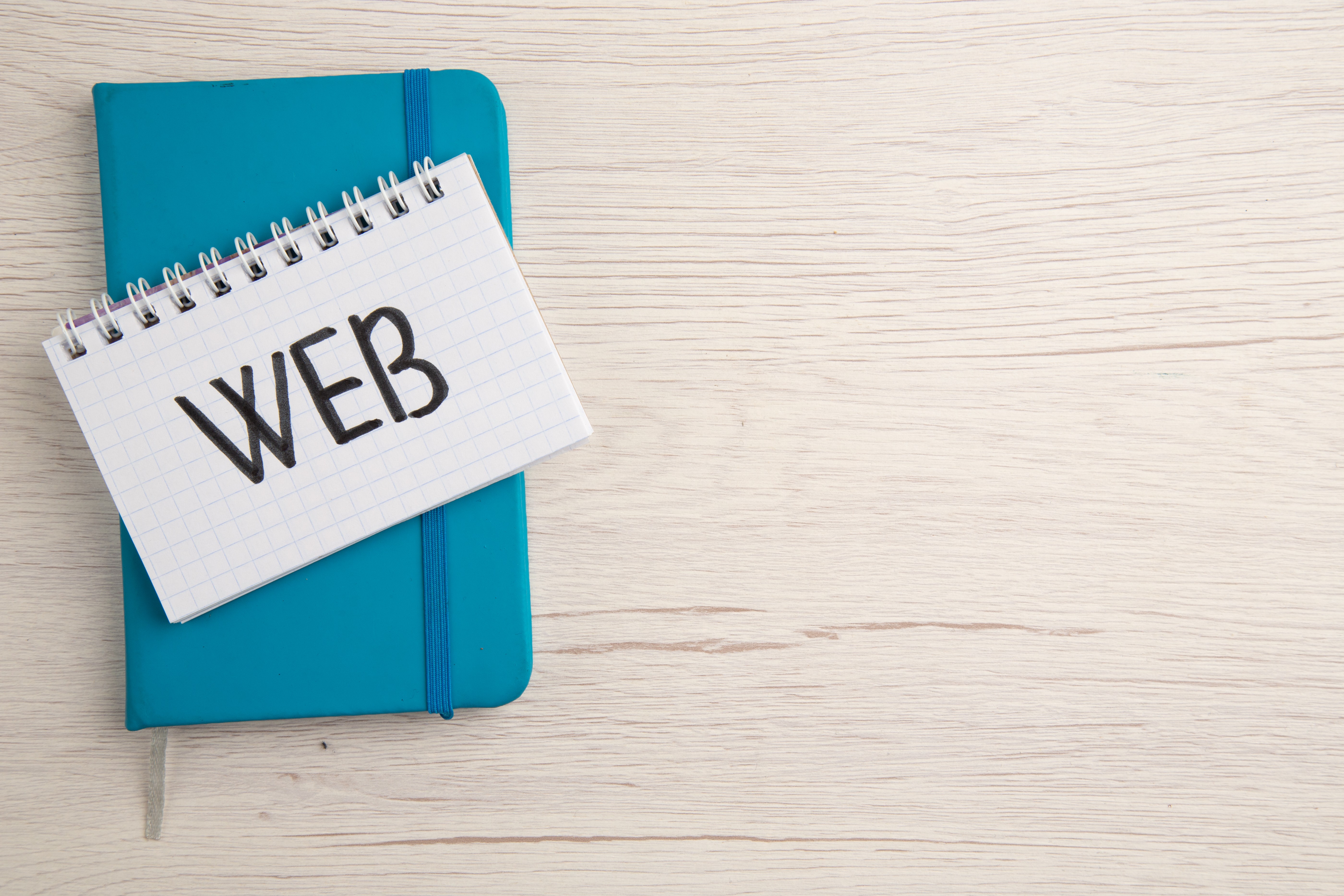 Top view of a notepad with the word ‘WEB’ written on it, placed over a blue diary on a light wooden desk — representing web design or digital planning