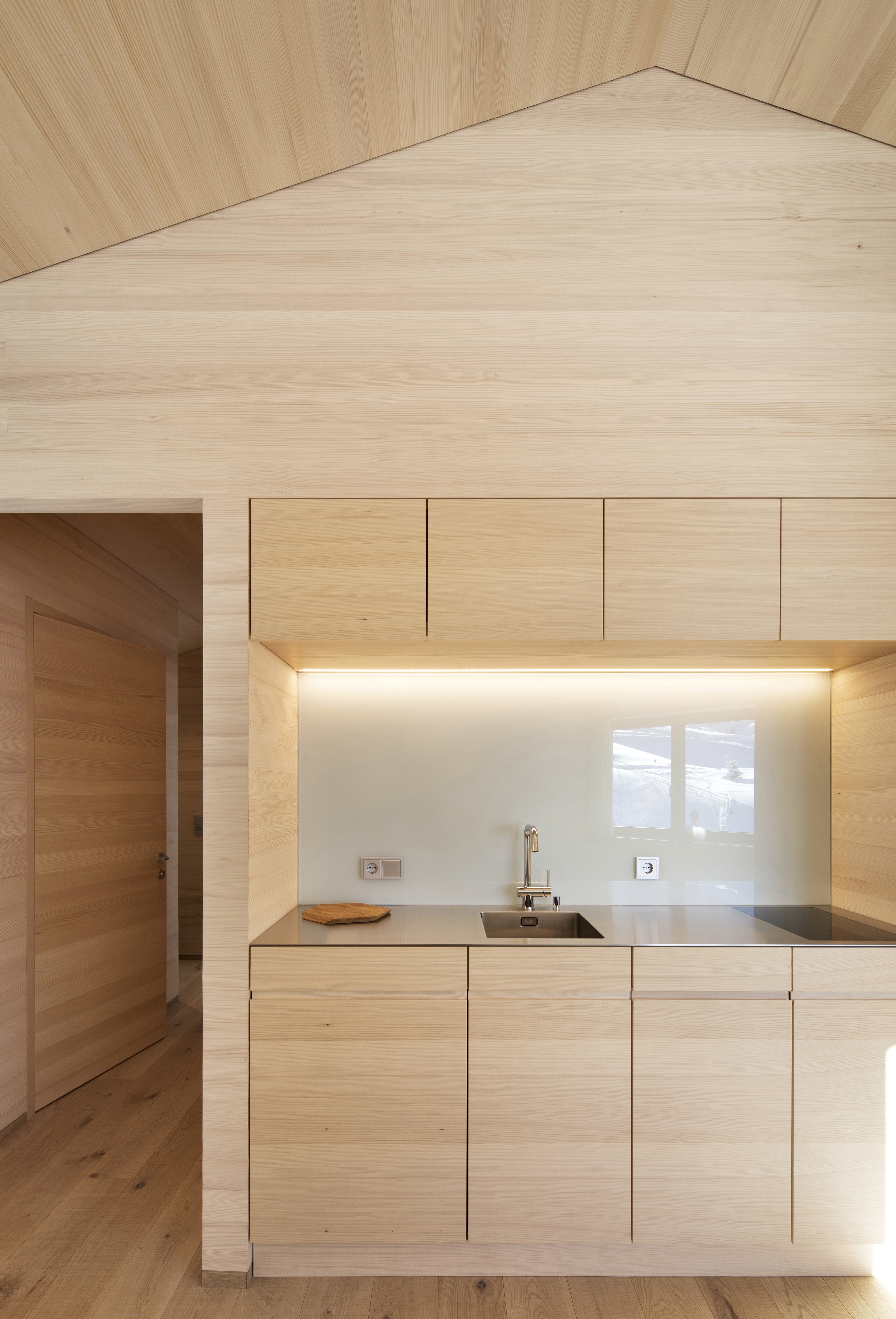 Compact kitchen in light natural timber with clean lines, integrated cabinets, a stainless-steel sink, and soft under-cabinet lighting