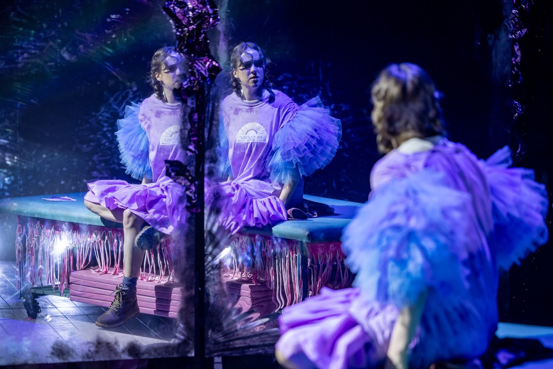 A performer in a lavender dress with ruffled sleeves sits on a bench and looks into a fractured mirror, their reflection appearing multiple times in the shimmering surface under atmospheric blue lighting.