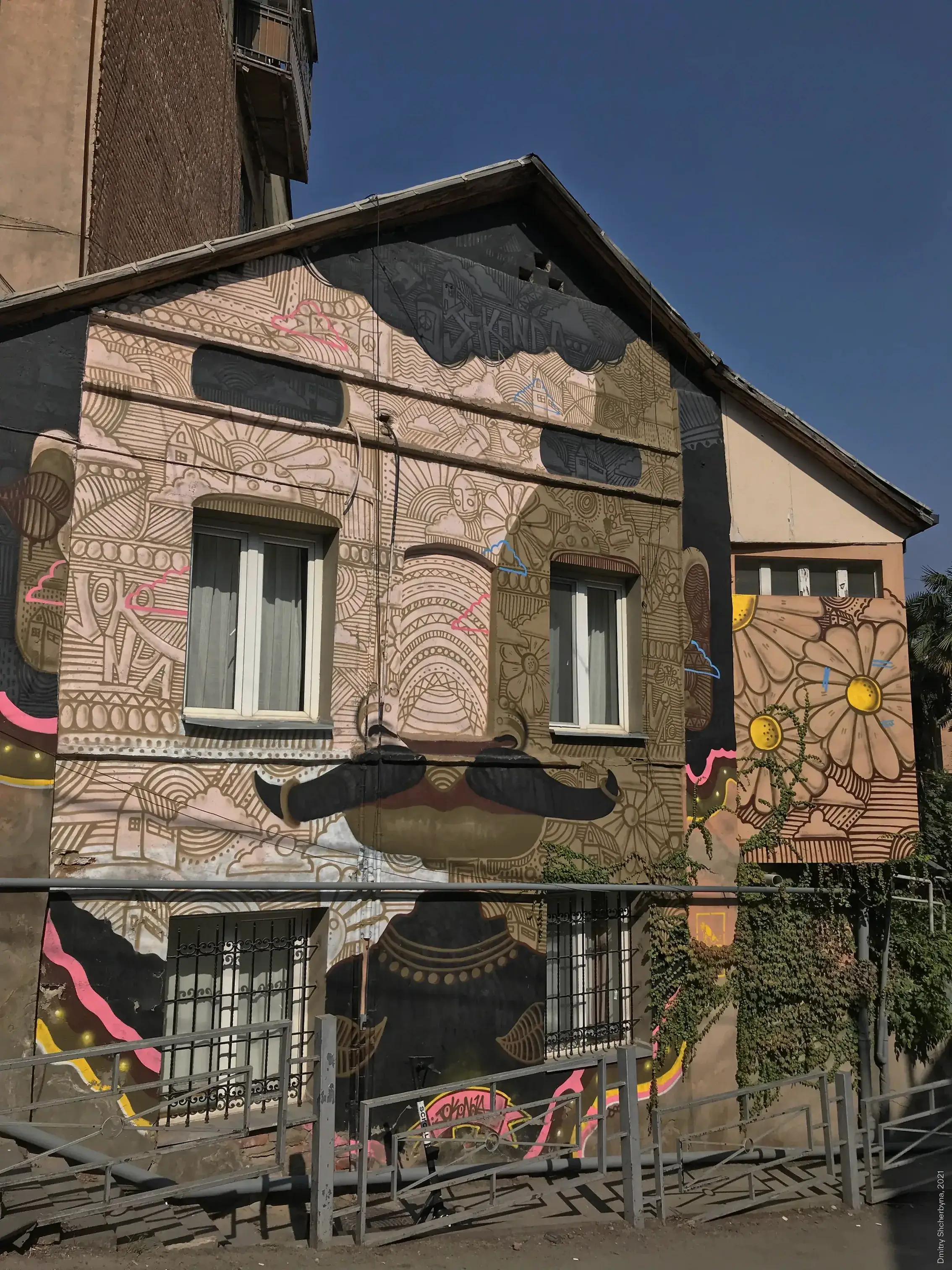 A mural covering the entire house, using the windows as the eyes of a large, hidden face composed of brown tan and walls filled with geometric and floral designs in pink, gold, and dark brown.
