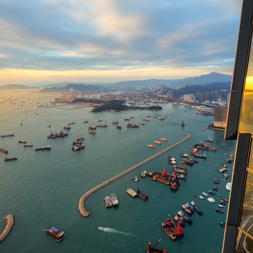 Felhőkkel borított ég alatt, egy nyüzsgő kikötő a levegőből nézve, számos hajóval és csónakkal, amelyet városkép és hegyek vesznek körül.