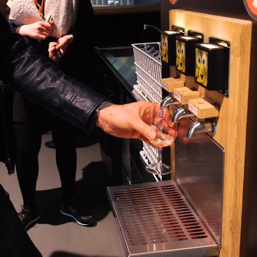 A person in a leather jacket fills a glass from a beer tap that has cartoon faces on the handles in a public setting.