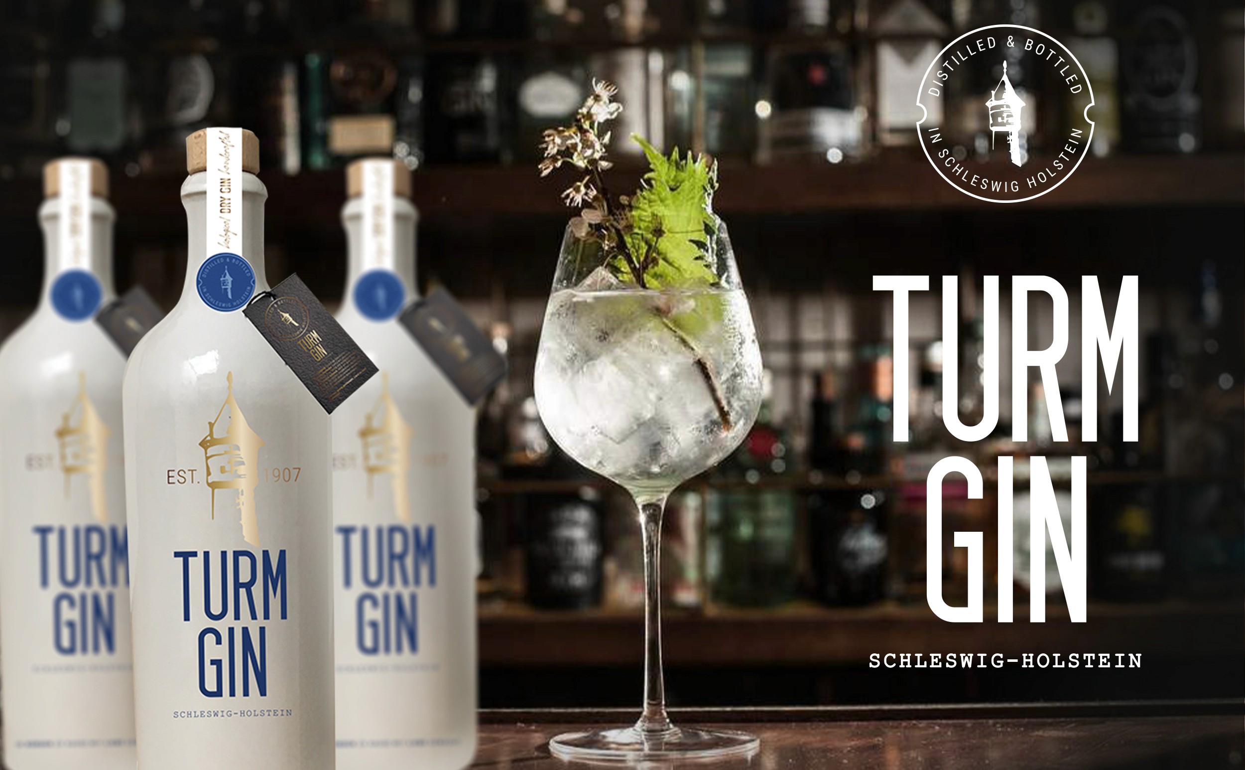Drei weiße Turmgin Flaschen mit der Aufschrift 'TURM GIN SCHLESWIG-HOLSTEIN' neben einem mit Eis und Kräutern gefüllten Gin-Glas auf einer Bar-Theke