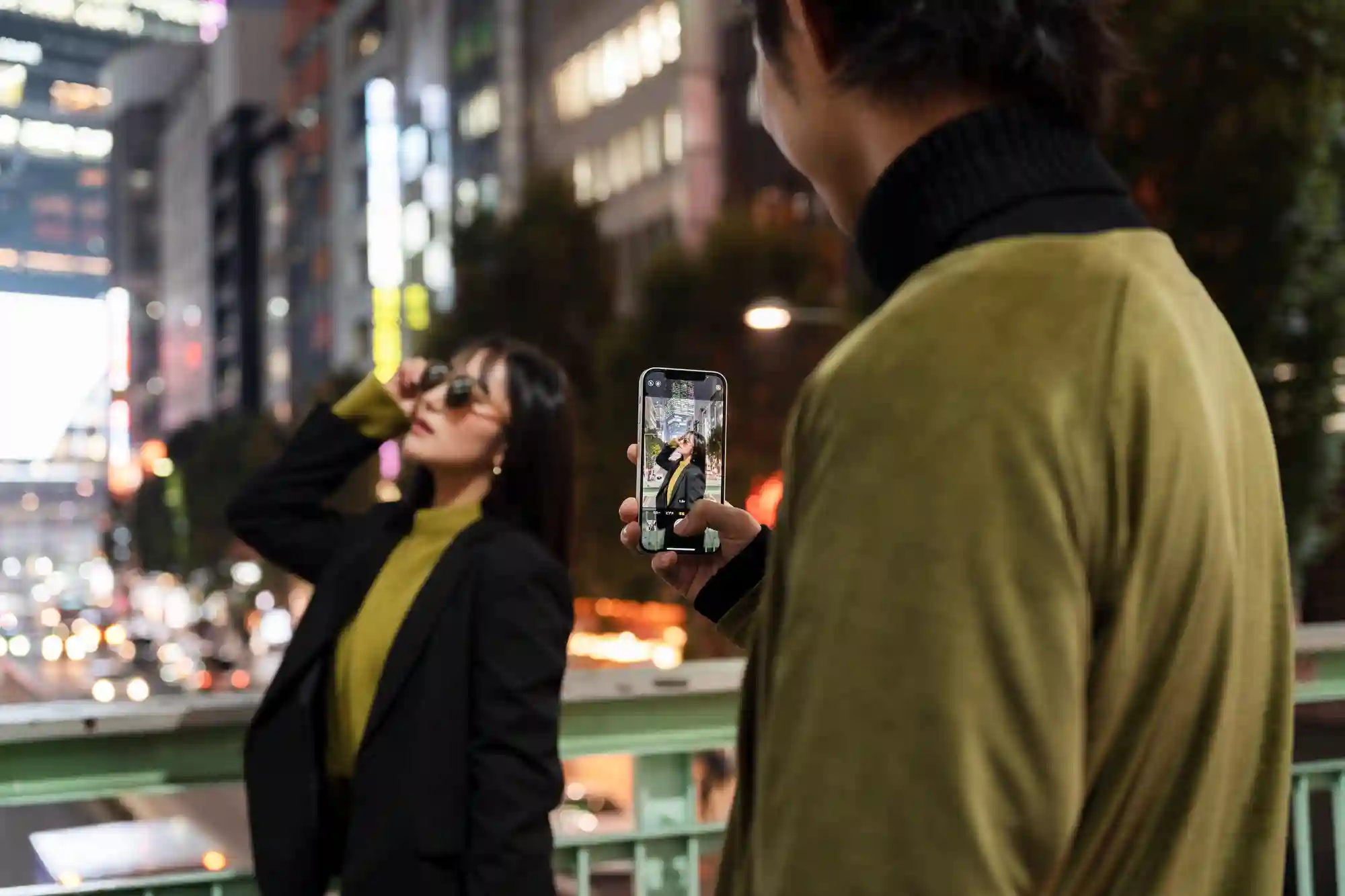 A man uses a smartphone to capture a stylish night portrait of a woman posing on a city bridge.