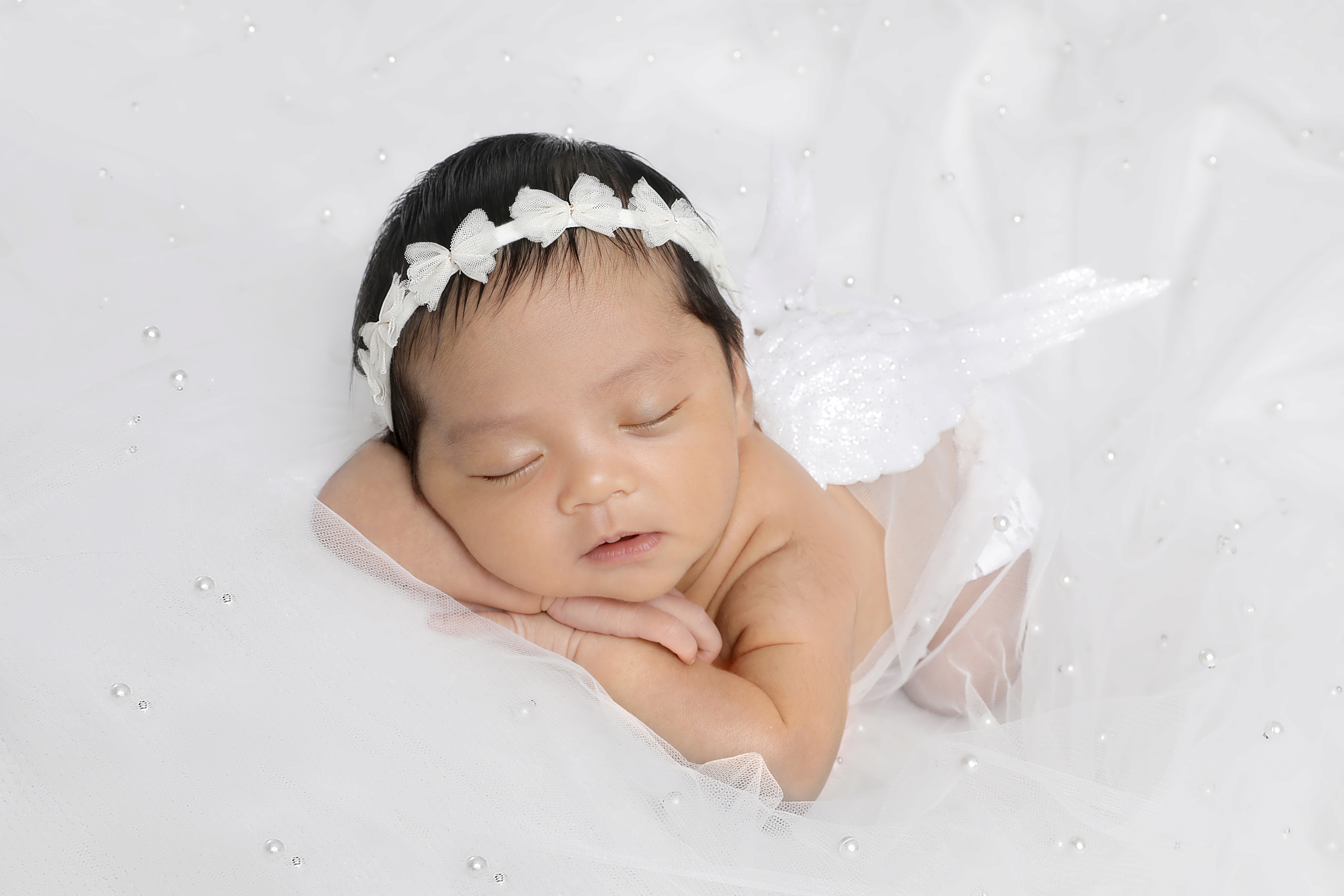 A peaceful sleeping newborn wears a white bow headband and angel wings on a sparkling white backdrop, captured with soft lighting during an infant photography shoot.