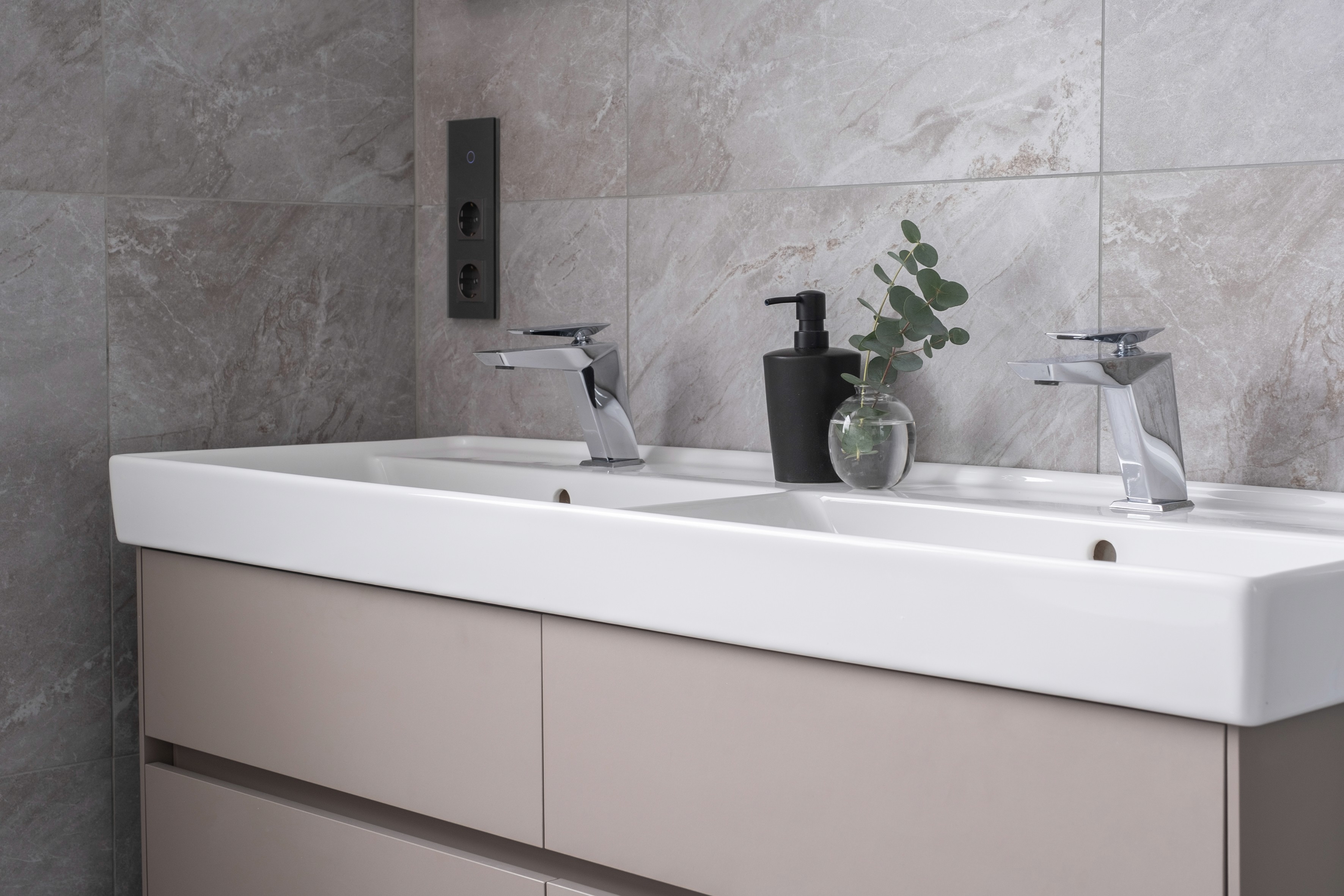 Double bathroom vanity with white rectangular basin, chrome waterfall taps, grey marble-effect tiles, and eucalyptus in glass vase