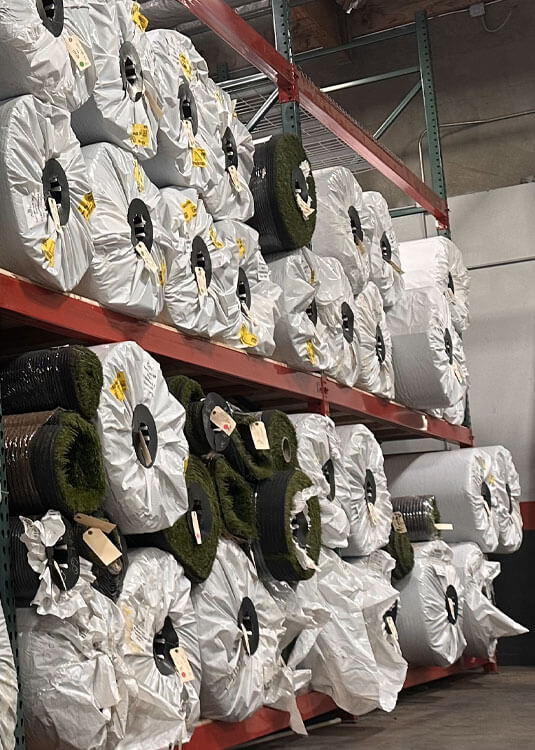 This expansive view of the AGL Grass warehouse showcases an abundance of supplies and neatly arranged rolls of artificial turf products such as Majestic 70, Saratoga 60, and Monte Carlo 60.