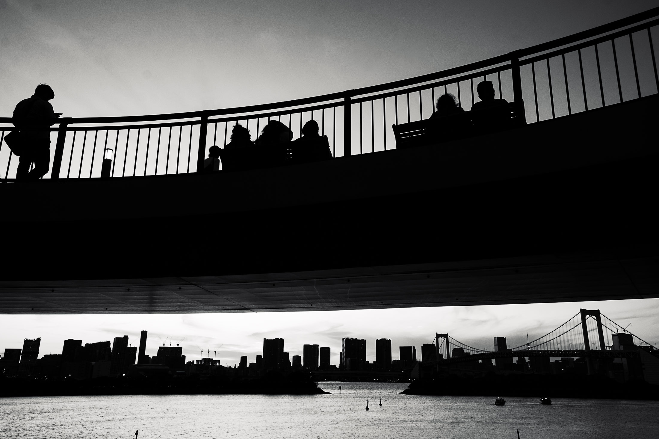 People sitting on a pedestrian bridge overlooking a city skyline and water, Tokio skyline black and white urban landscape photograph.