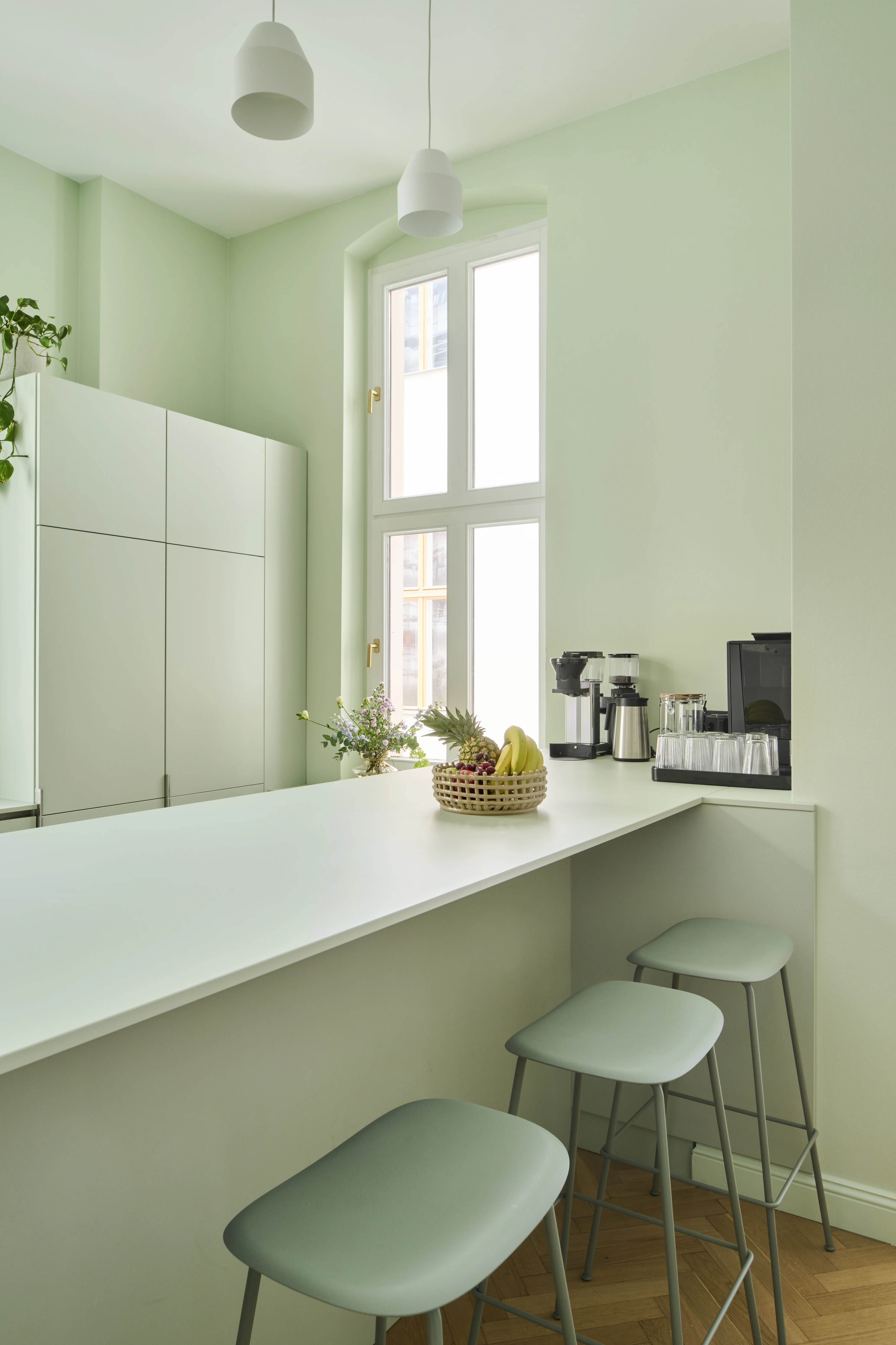 Mint-green kitchen at INTERMATE BERLIN with a long pale counter, sage bar stools, coffee machines and thermos flasks, a fruit basket and white pendant lights by a tall window.