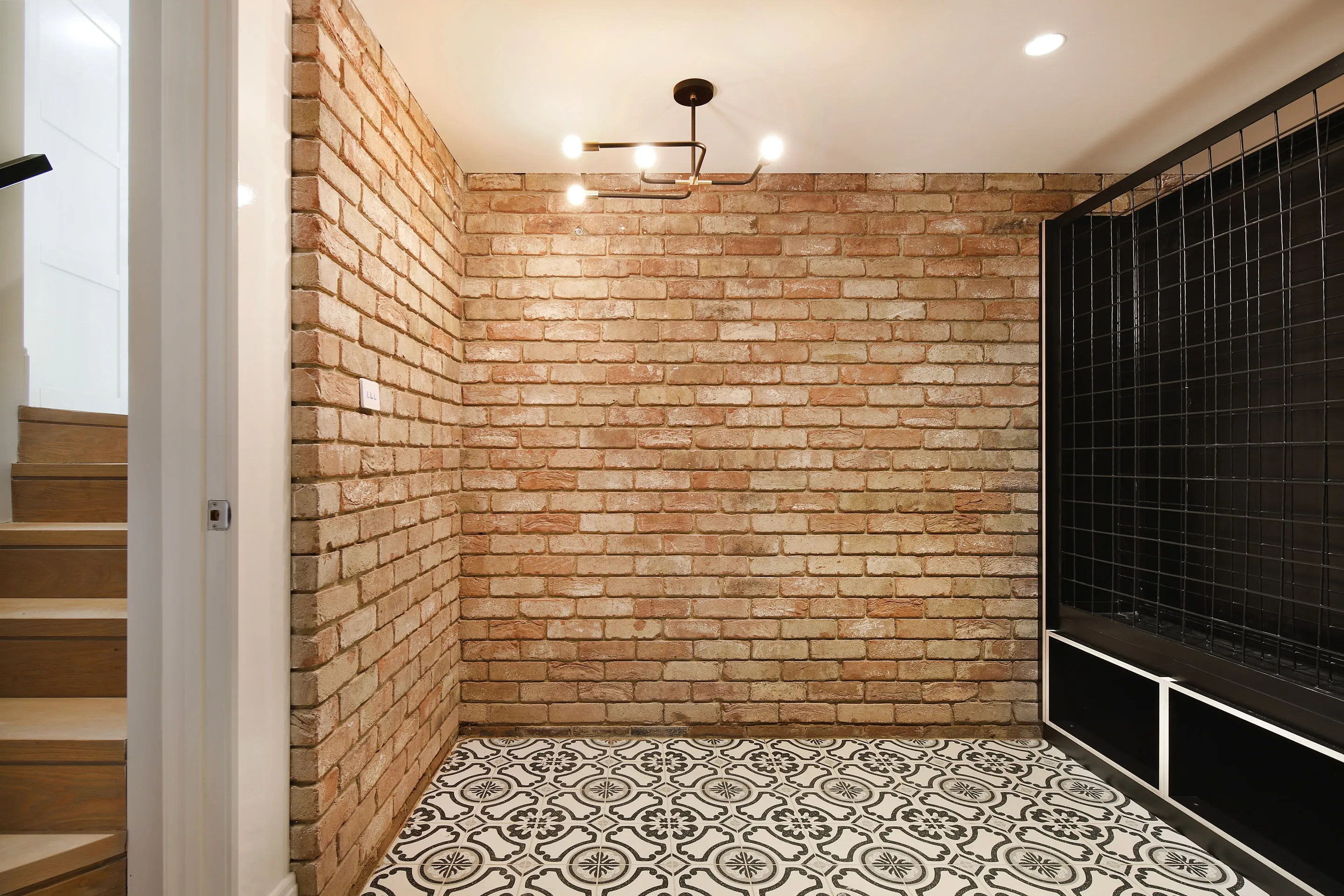 Recycled-brick cellar with patterned black-and-white floor tiles, a black steel-and-mesh wine wall with white storage cubes and a glimpse of the timber staircase