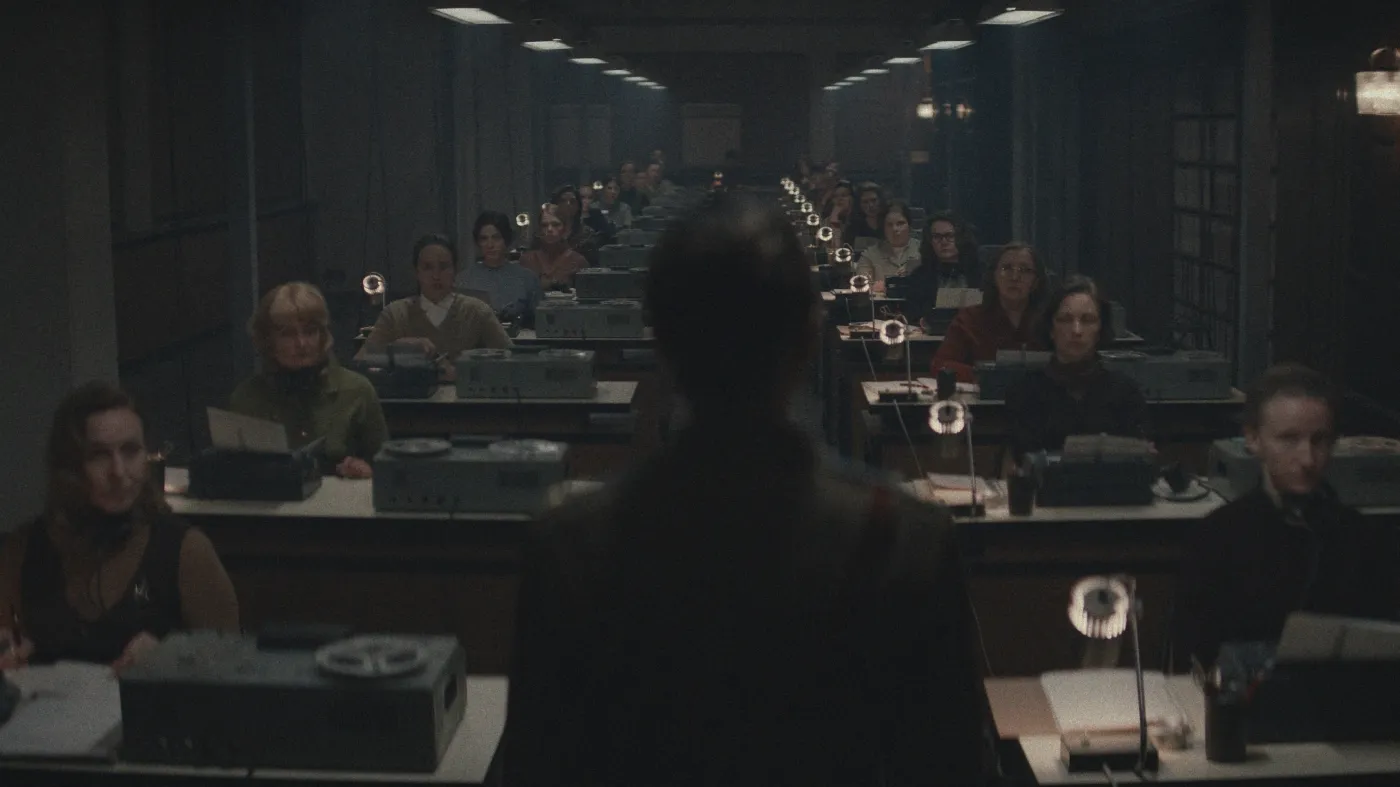 A dimly lit, crowded office room featuring rows of workers typing on vintage typewriters, with an indistinct shadowed figure standing at the front, creating an atmosphere reminiscent of a 1940s typing pool.