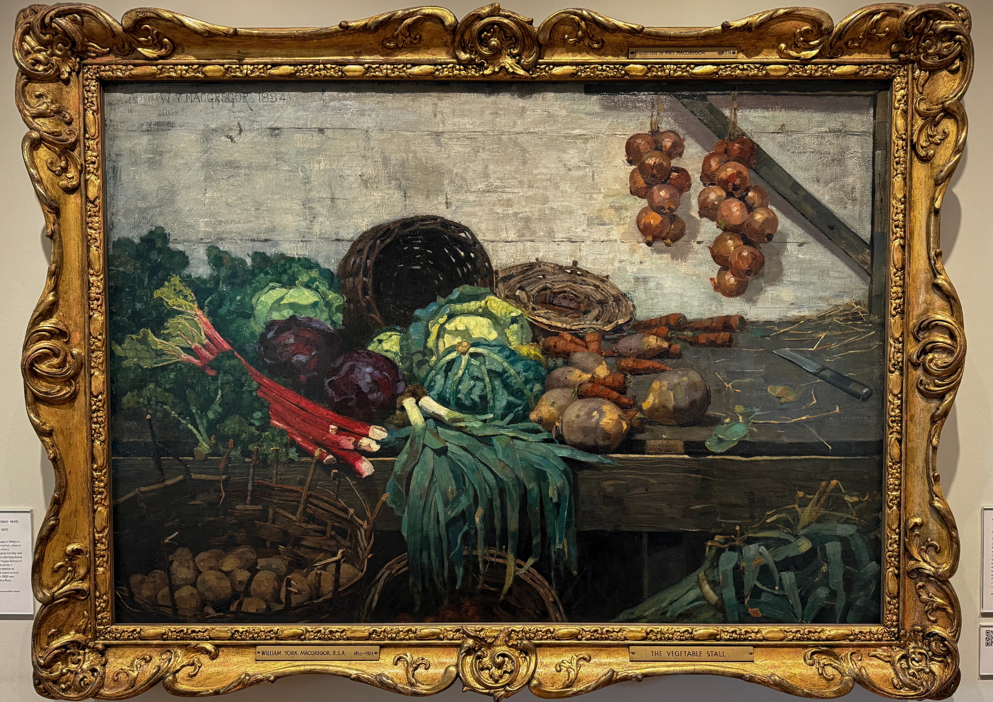 Painting of a market stall piled with vegetables: red-stemmed chard, cabbages, a dark basket, onions hanging above, in muted earthy tones.