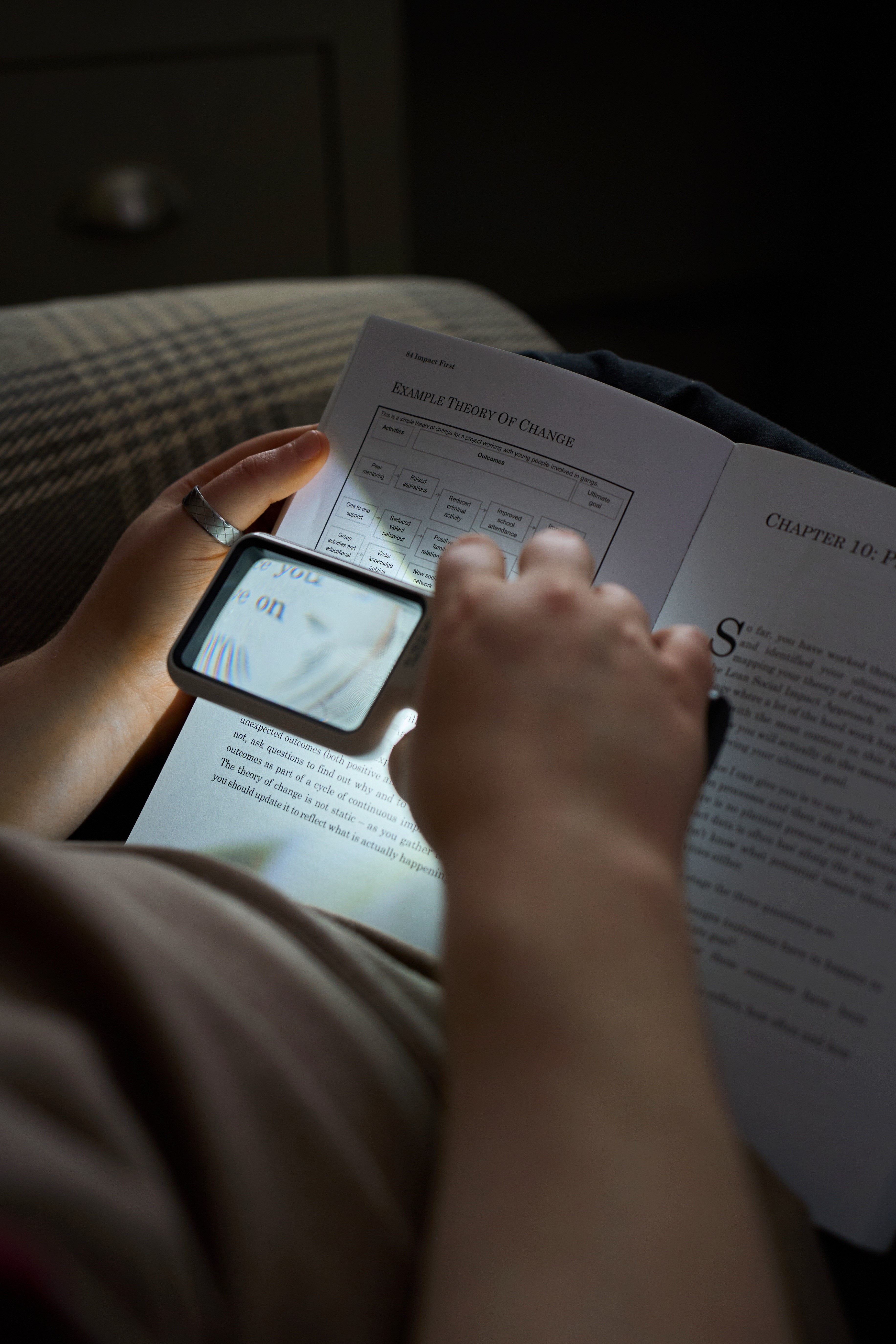 Close-up of a person holding an illuminated magnifier over an open book. The text on the page becomes larger and distorted through the lens, showing how someone with sight loss might need magnification to read. The section of the book visible includes charts and small printed text.