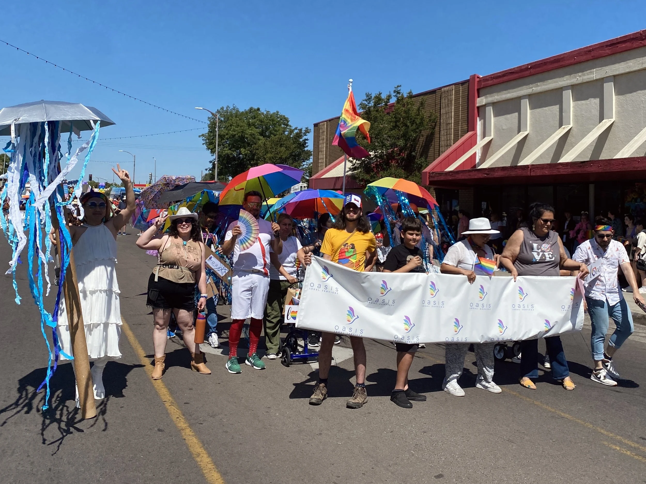 Uma cena vibrante de desfile mostra um grupo diverso de pessoas marchando alegremente por uma rua, algumas segurando guarda-chuvas e faixas com as cores do arco-íris, enquanto um participante balança fitas presas a um bastão alto, celebrando sob um céu azul e limpo.