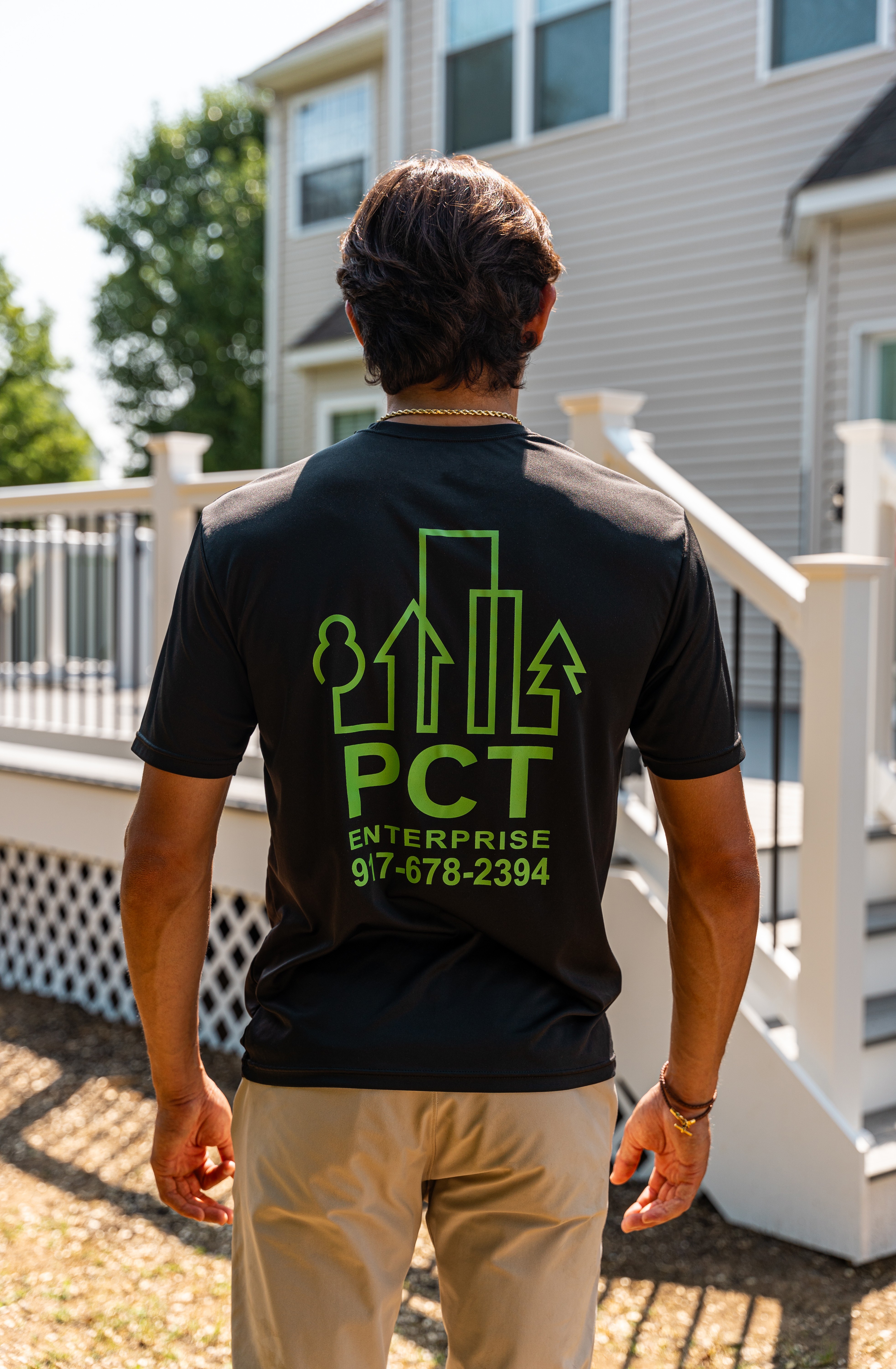 A person wearing a black t-shirt with green graphics and text stands outside a house with a deck and white railing, surrounded by trees under a sunny sky.