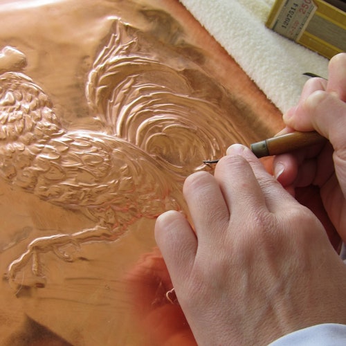 Tay đang dùng công cụ để khắc họa tiết con gà trên tấm đồng. Bên cạnh tấm đồng là một hộp dụng cụ.