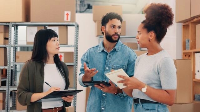 Três pessoas conversando em um armazém; caixas de papelão organizadas em prateleiras metálicas ao fundo.