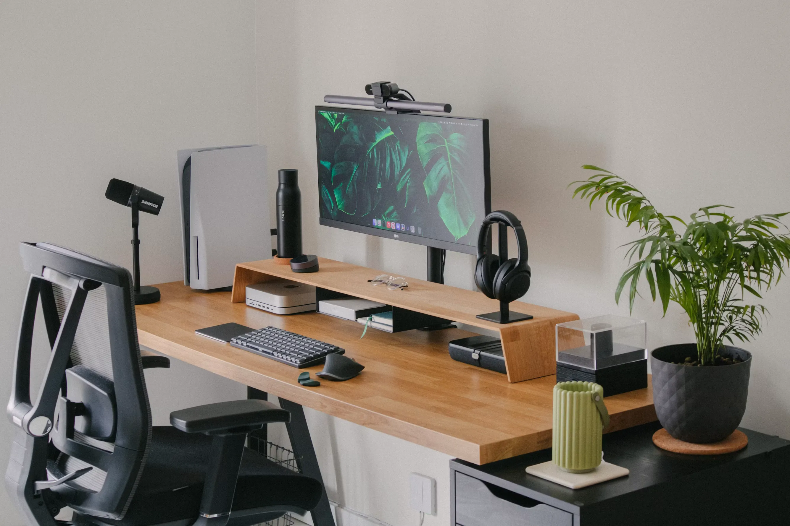 A modern and organized home office desk setup with a minimalist wooden desk and matching monitor riser. The workspace features an ultrawide monitor with a screen light bar, a Mac Mini, a PlayStation 5, a mechanical keyboard, an ergonomic mouse, a professional microphone, and black headphones on a stand. A sleek office chair sits to the left, and a vibrant green potted plant sits on an adjacent black filing cabinet against a plain white wall.