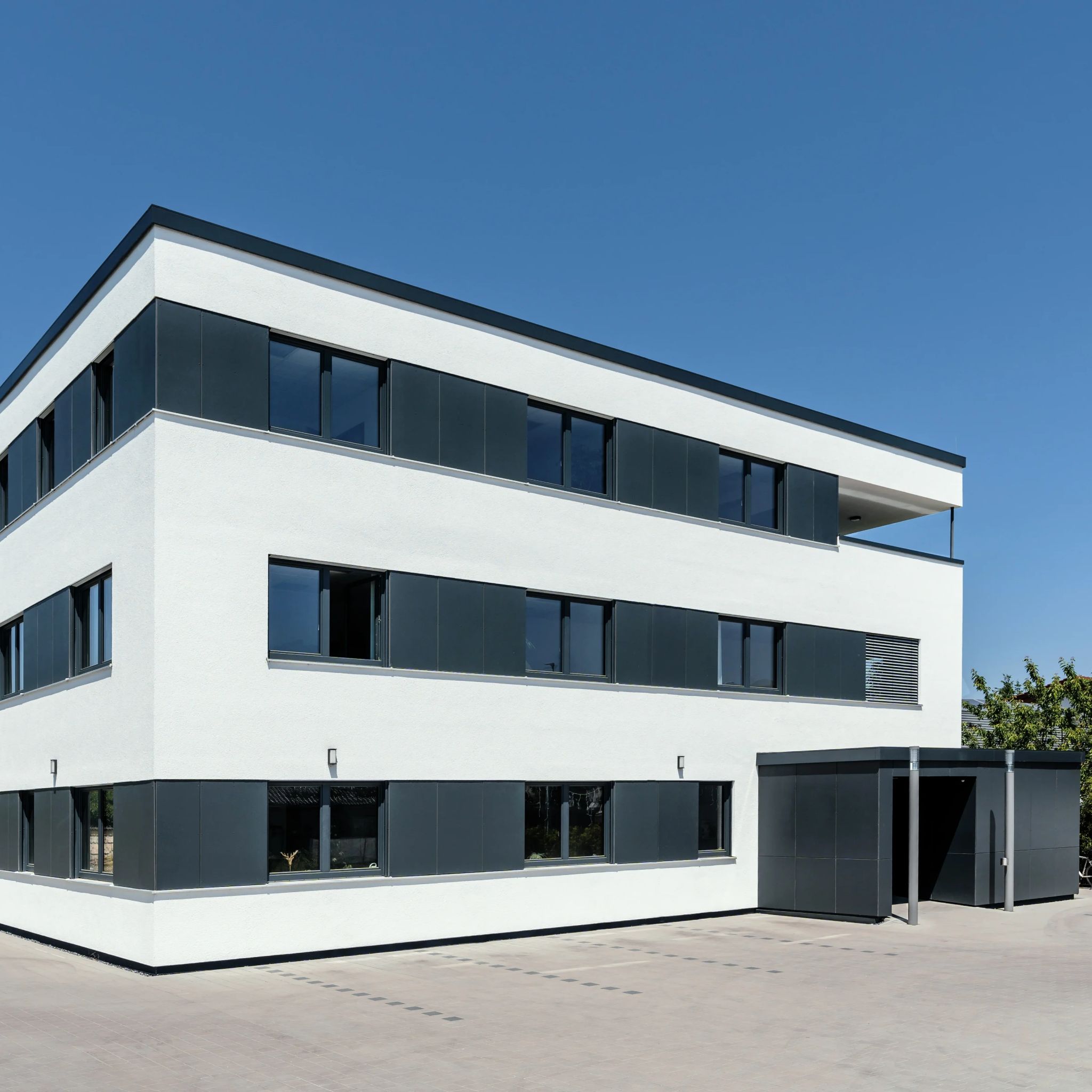 Modernes Bürogebäude mit weißer Putzfassade, dunklen Fensterrahmen und schiefergrauen Verkleidungen unter blauem Himmel.